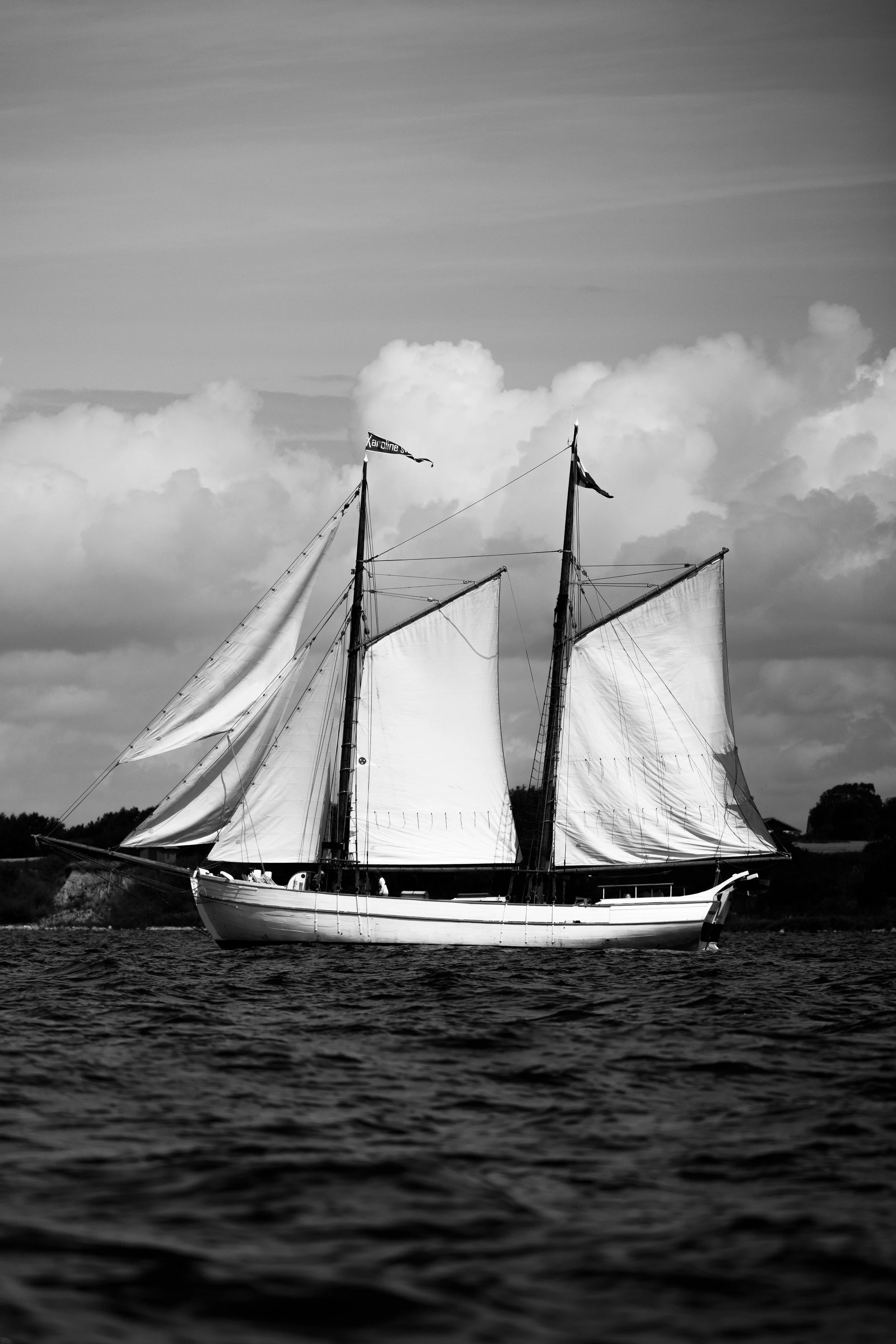 A black-and-white photo of a sailing ship with multiple masts and sails on the water, with a cloudy sky in the background.