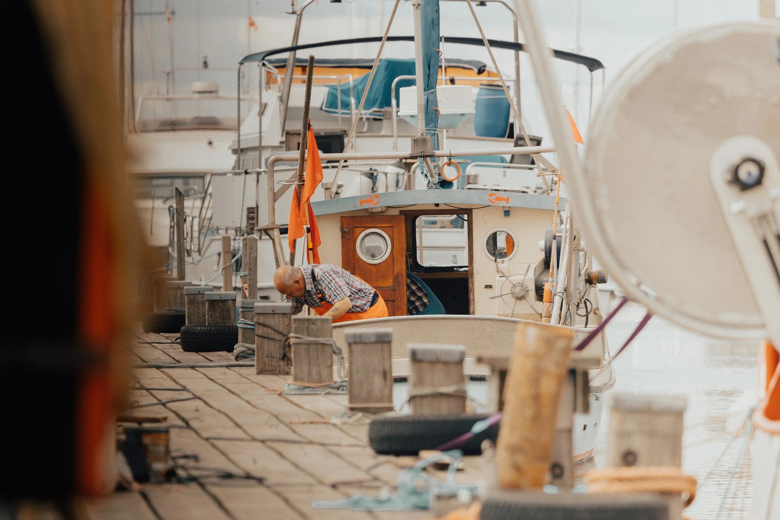 An elderly man wearing an orange apron and plaid shirt leaning over a wooden dock, working on a boat docked at a marina with other boats in the background.