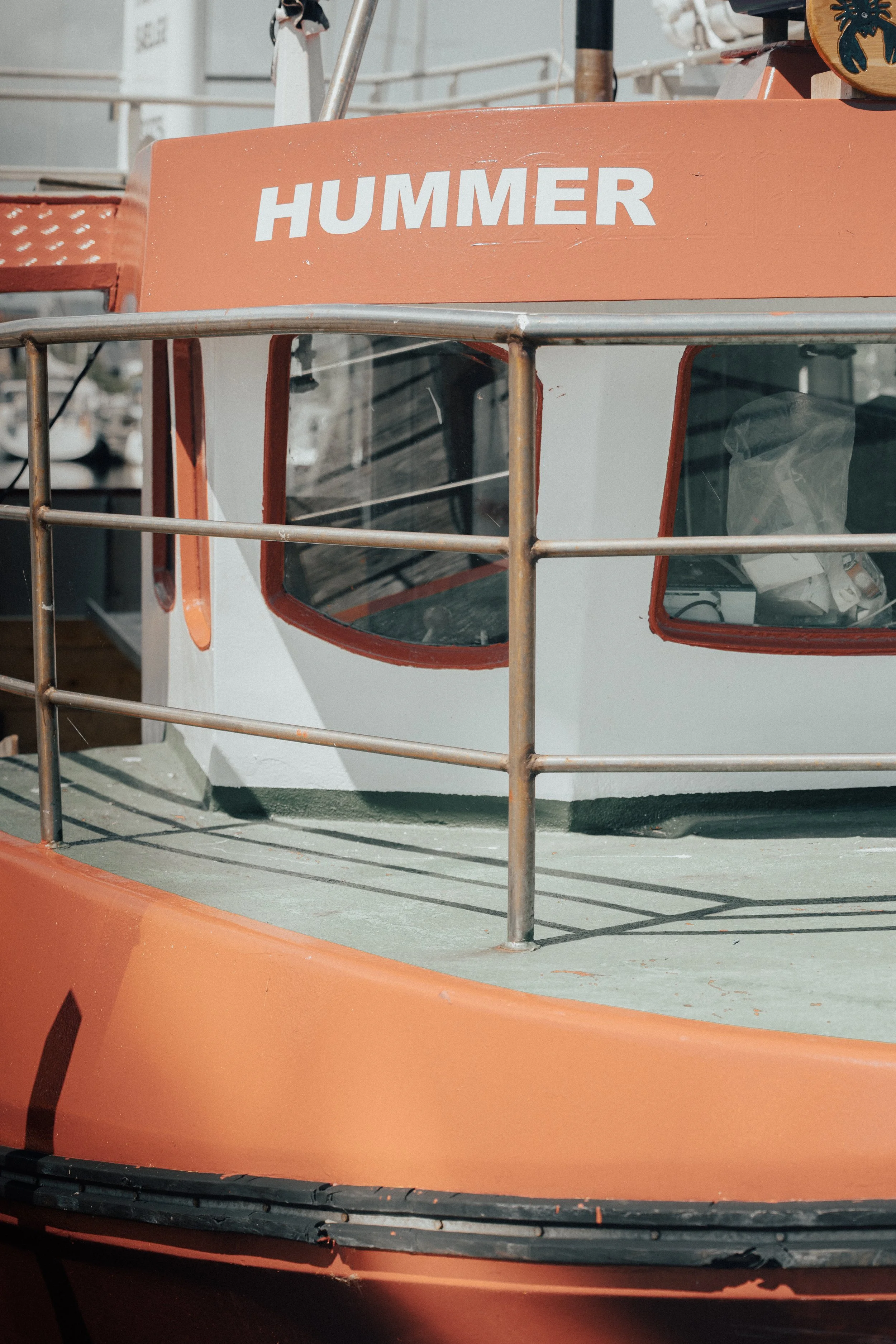 Close-up of the front section of a boat with a sign reading 'HUMMER' and metal railings. The boat is orange and white with windows and some equipment inside.