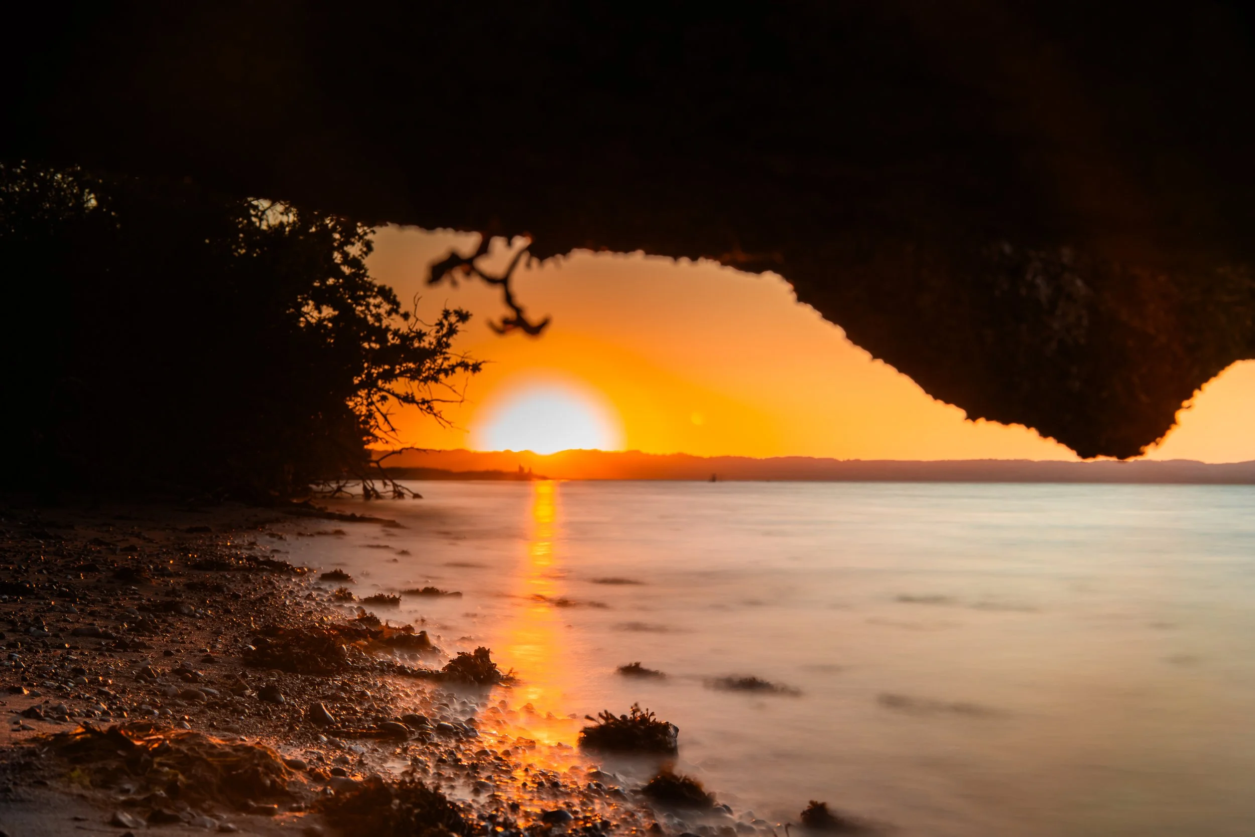 Sunset view through a rocky overhang, with silhouettes of trees on the shore and calm water reflecting the orange sky.