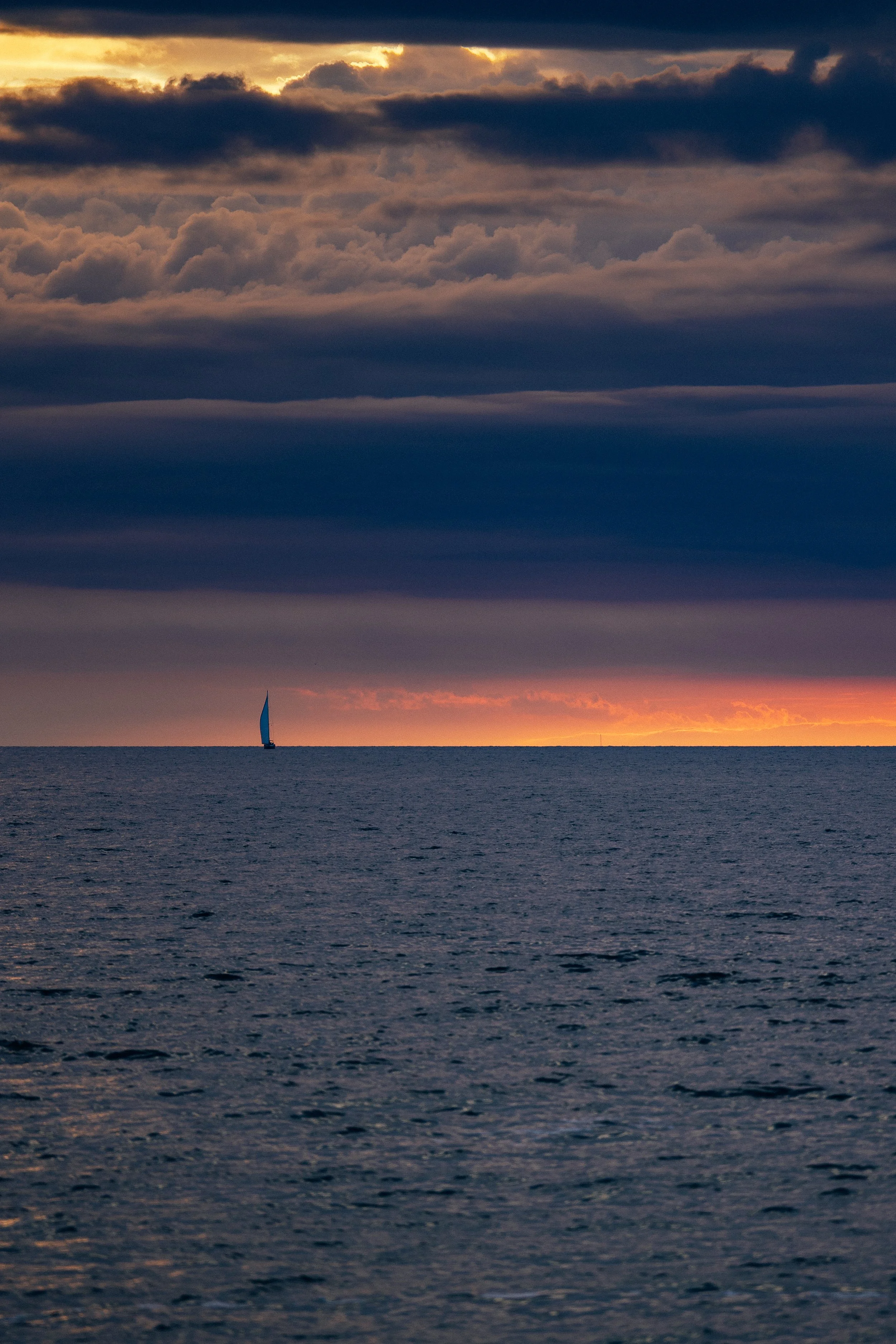 A sailboat with a blue sail on calm ocean waters during sunset, with a colorful sky filled with layered clouds.