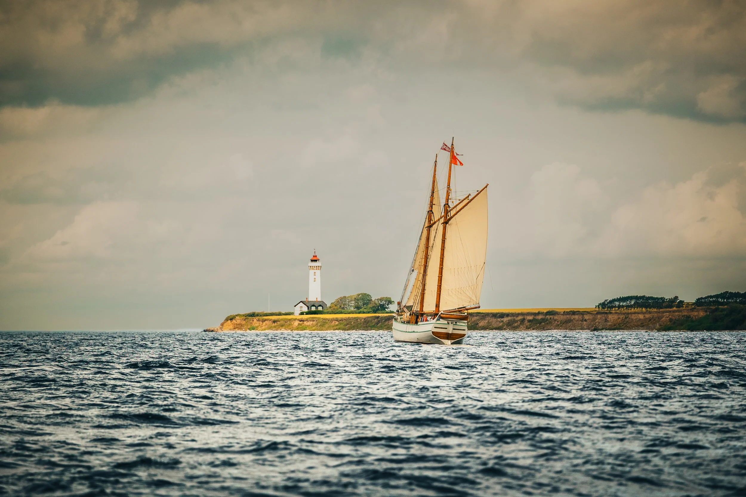 Sailing ship on water near a lighthouse on a distant coastline, under a cloudy sky.