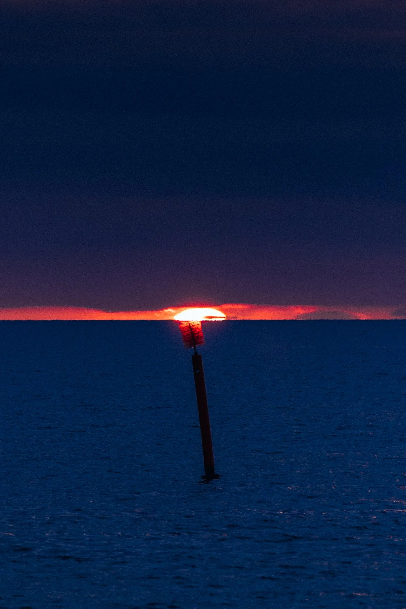 A sunset over the ocean with a broken navigation marker leaning at an angle in the water.