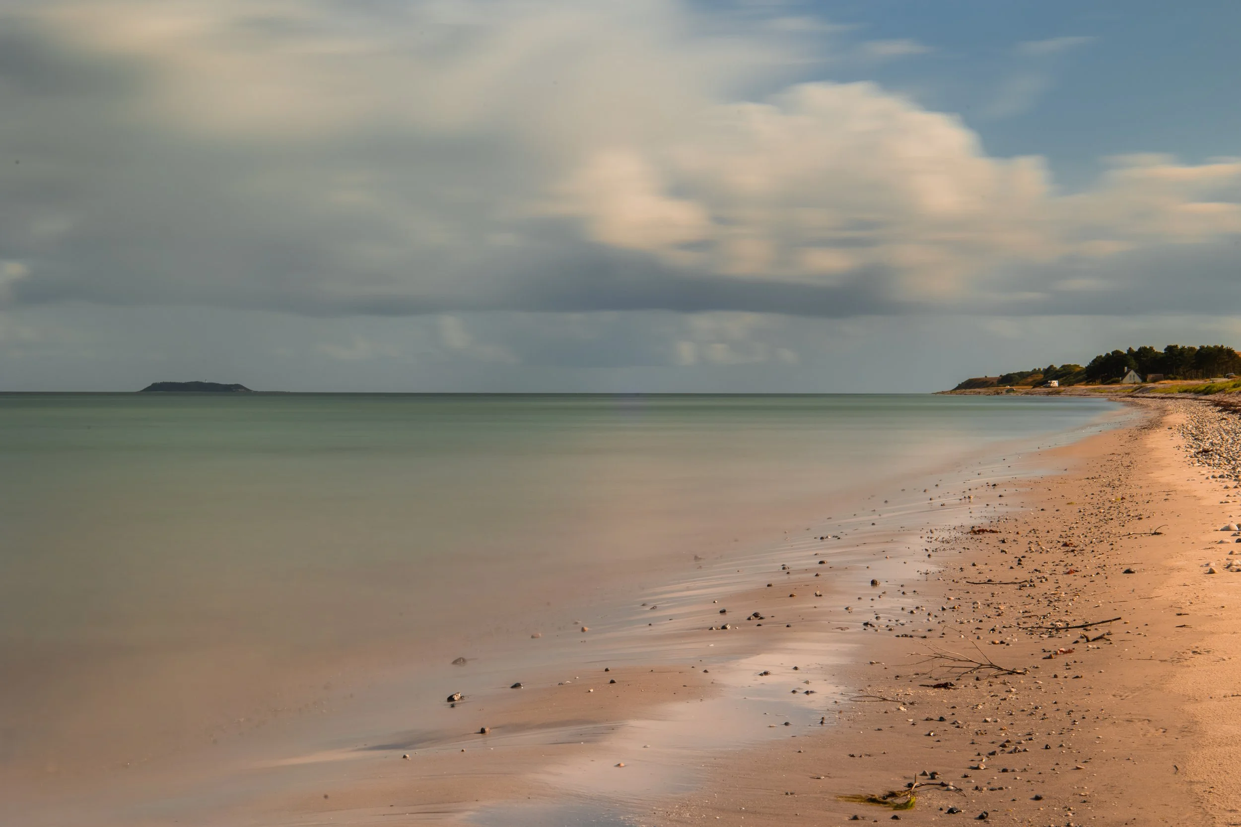 A serene beach scene with smooth sand, small rocks, and driftwood, calm ocean water, and a partly cloudy sky, with a distant island on the horizon and houses on the distant shoreline.