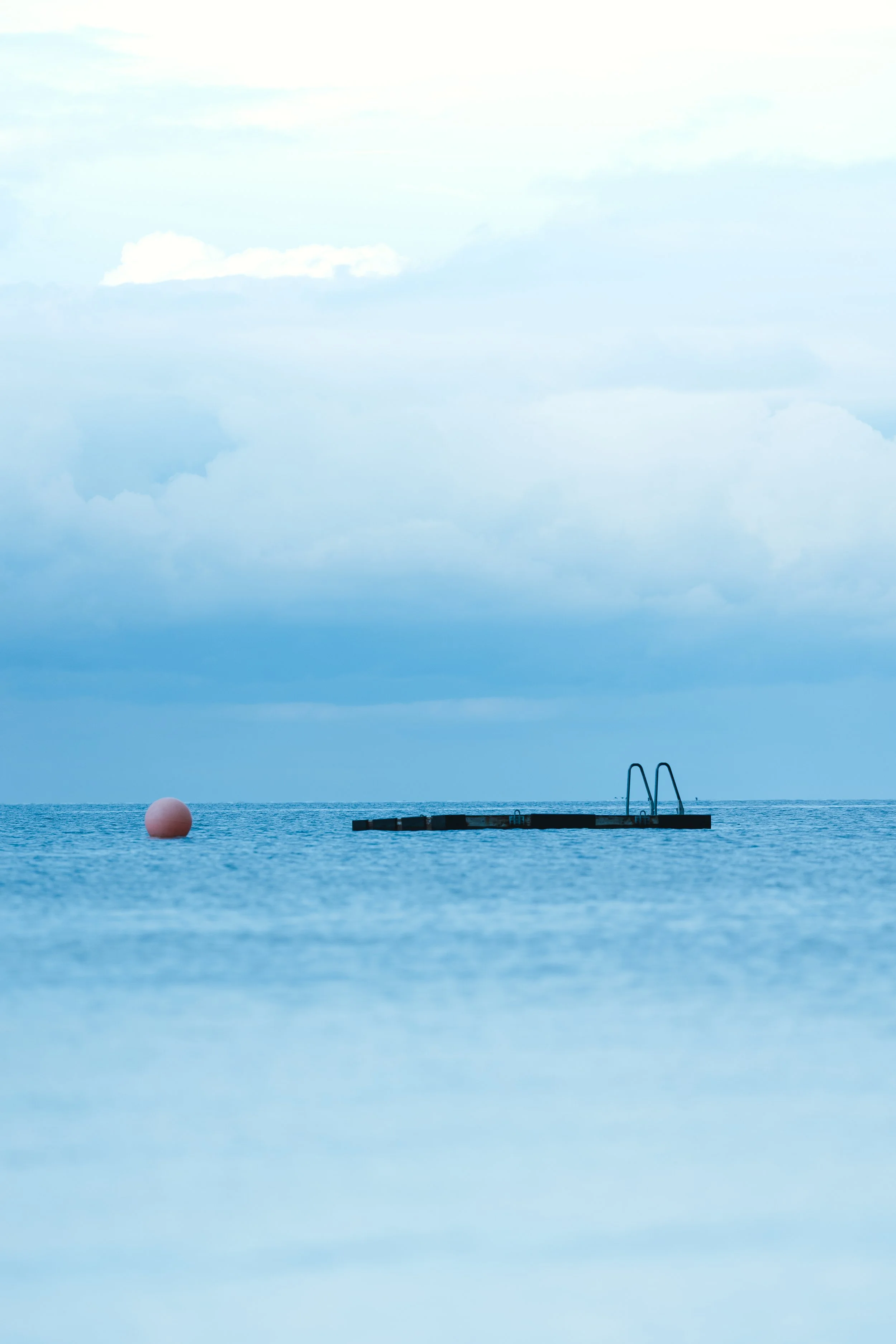 An overcast sky above a calm ocean with a floating dock, a structure with stairs, and a pink buoy nearby.