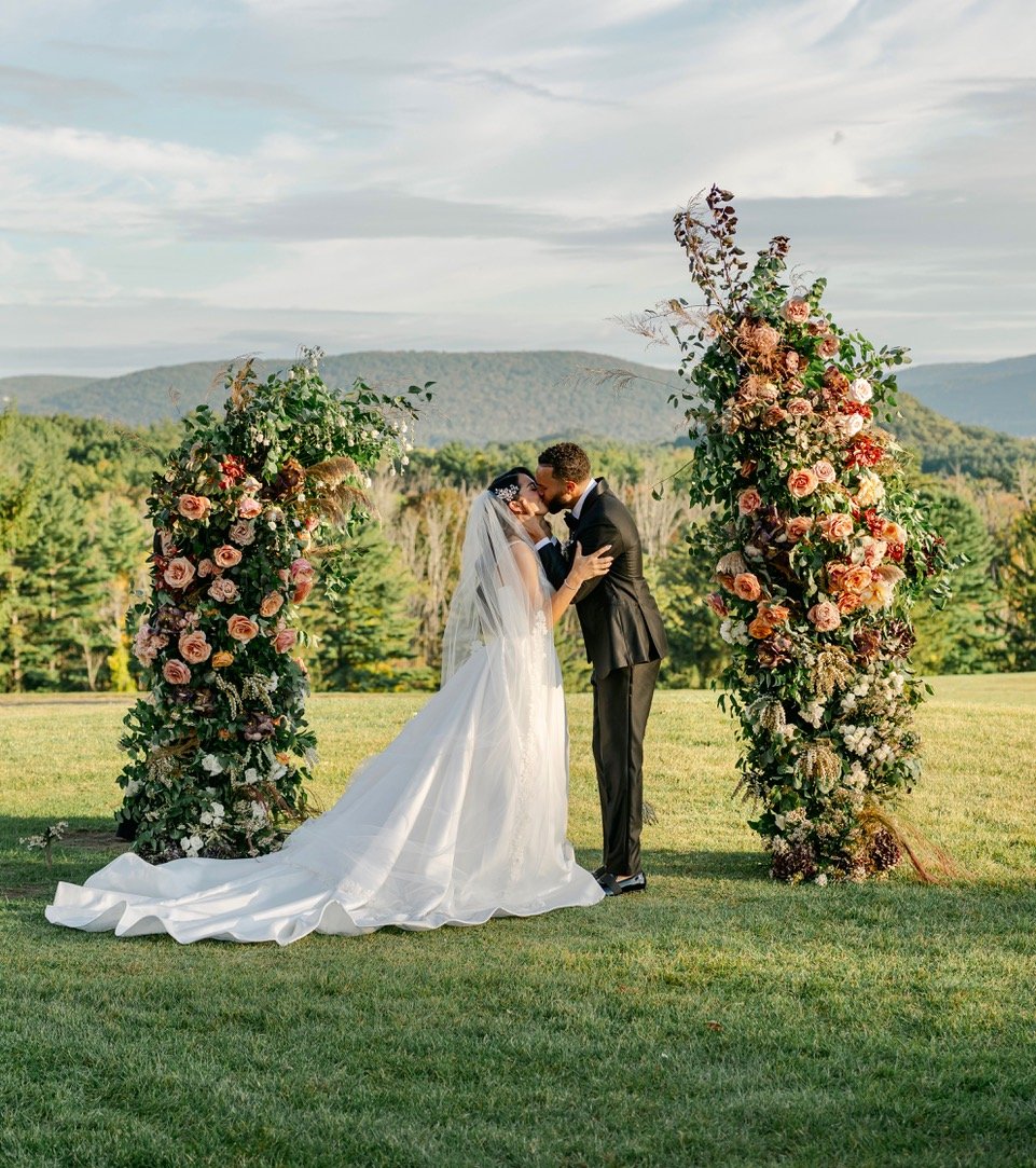 Bride and groom kissing under a floral arch at an outdoor wedding ceremony with a scenic mountain backdrop.
