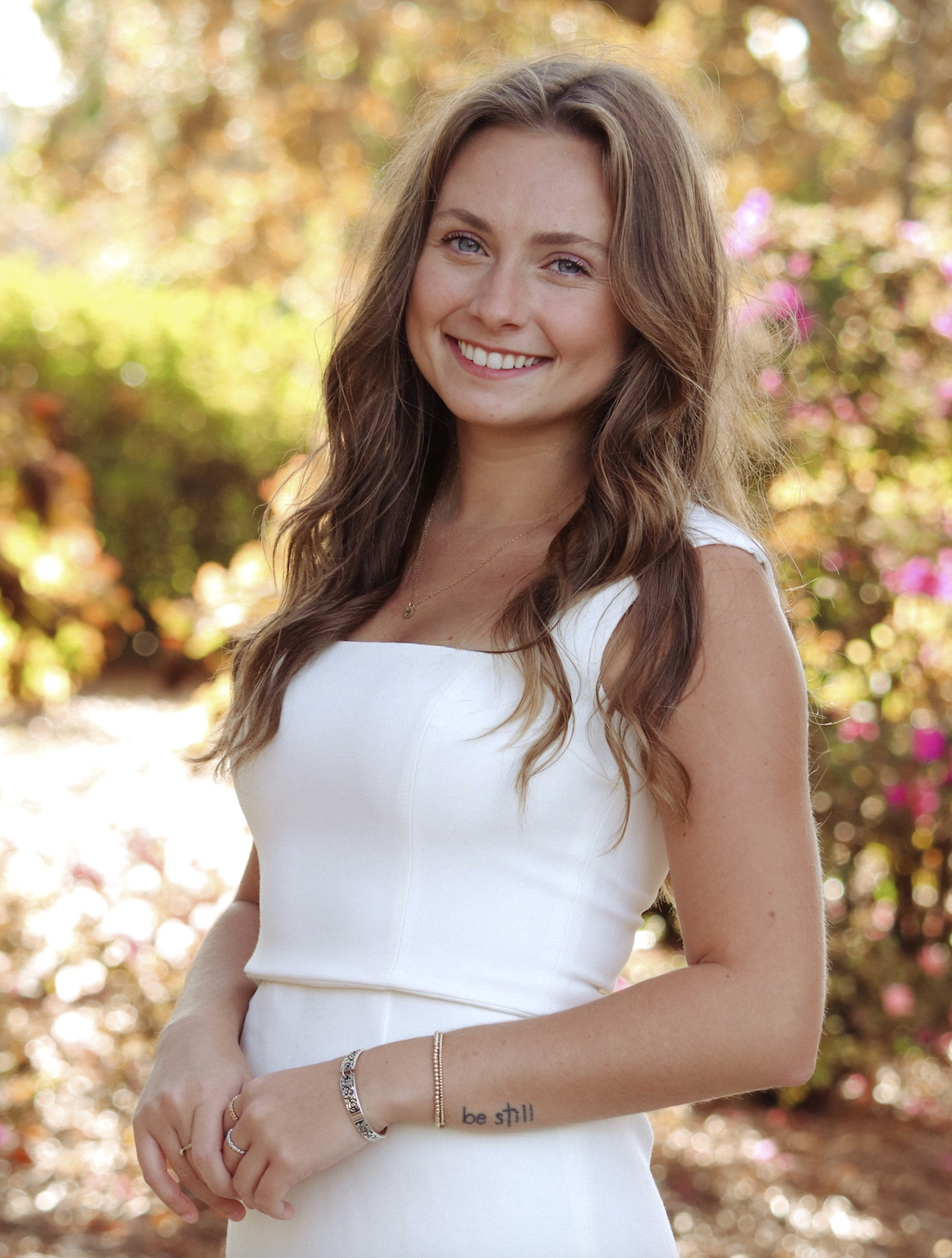 A young woman with long wavy brown hair, smiling and wearing a white sleeveless dress, standing outdoors with colorful autumn foliage in the background.