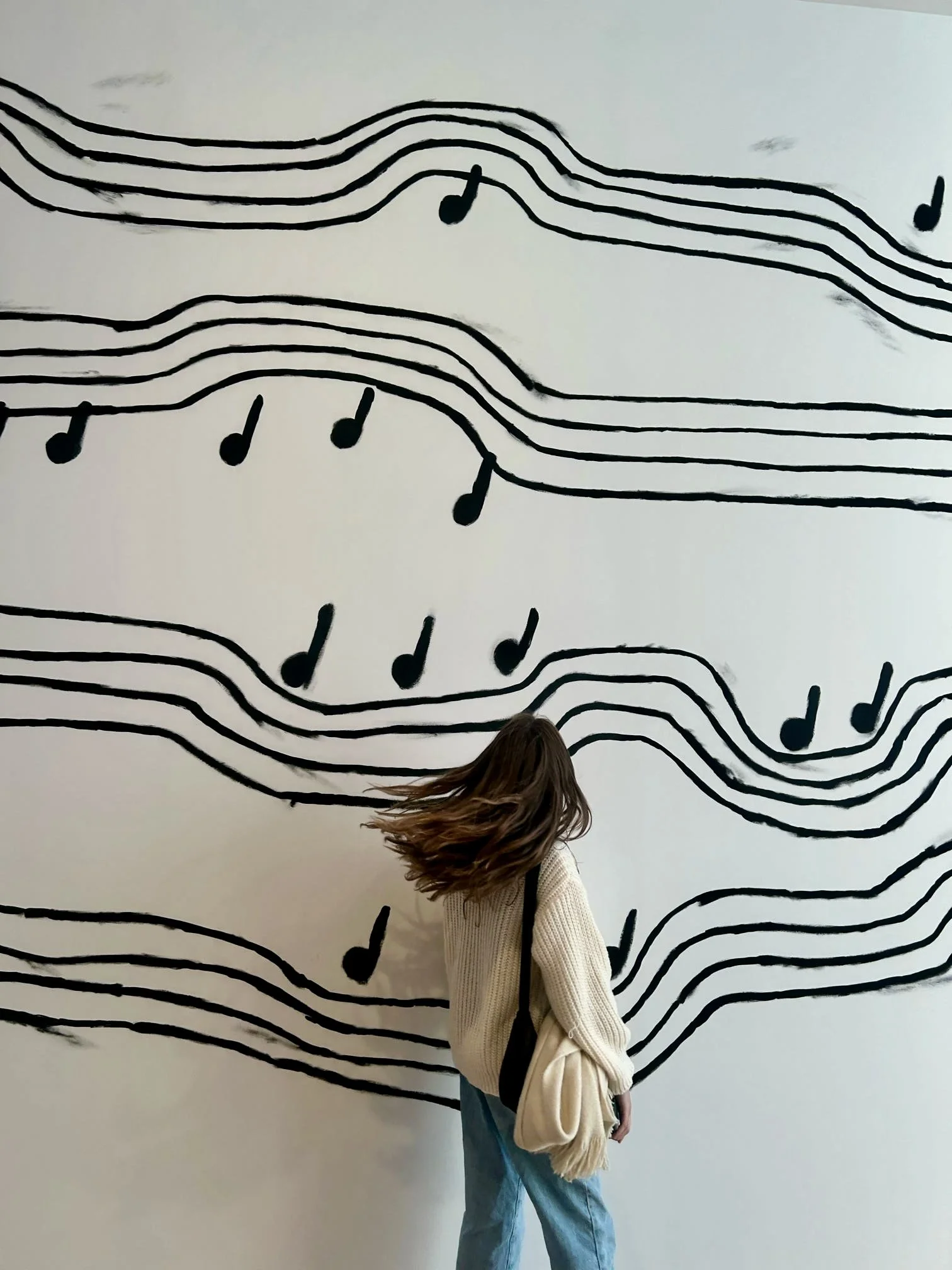 Woman with brown hair wearing a cream-colored sweater and jeans, standing in front of a white wall with black musical staff lines and notes painted on it.