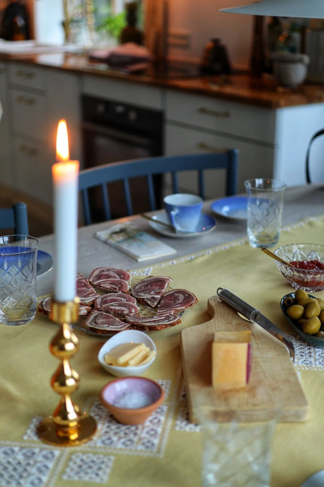 Table with candle, sliced meat, cheese on a board, olives, butter, salt, and glassware in a cozy kitchen setting. A unique opportunity for tourists to taste traditional Faroese cuisine.