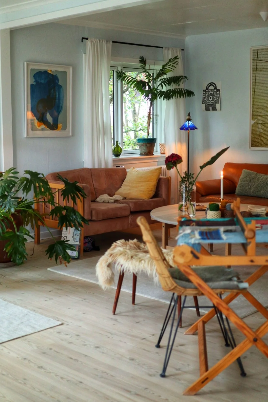 Cozy living room with brown leather couches, houseplants, a blue floor lamp, abstract wall art, wooden furniture, a round coffee table, a woven chair, and a large window with white curtains.