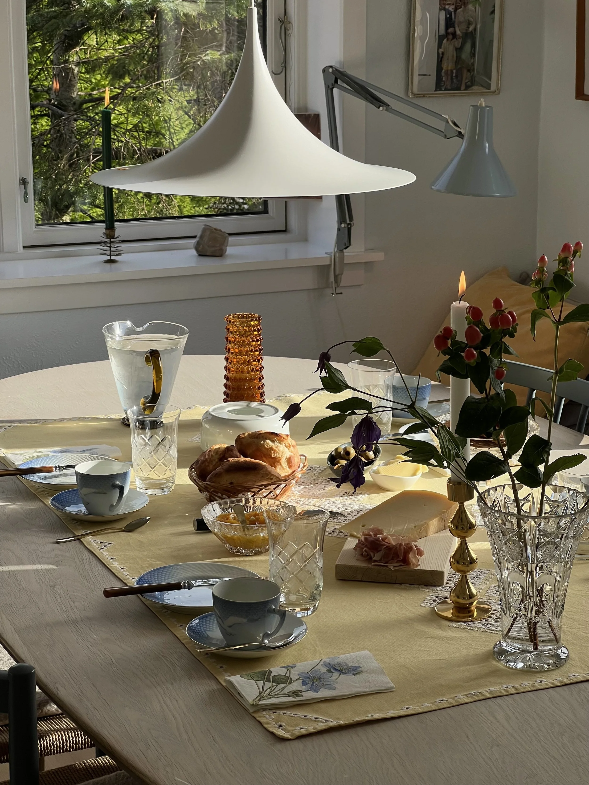 Table set for breakfast with cups, saucers, a vase with flowers, a candle, bread, and a pitcher of water near a window.