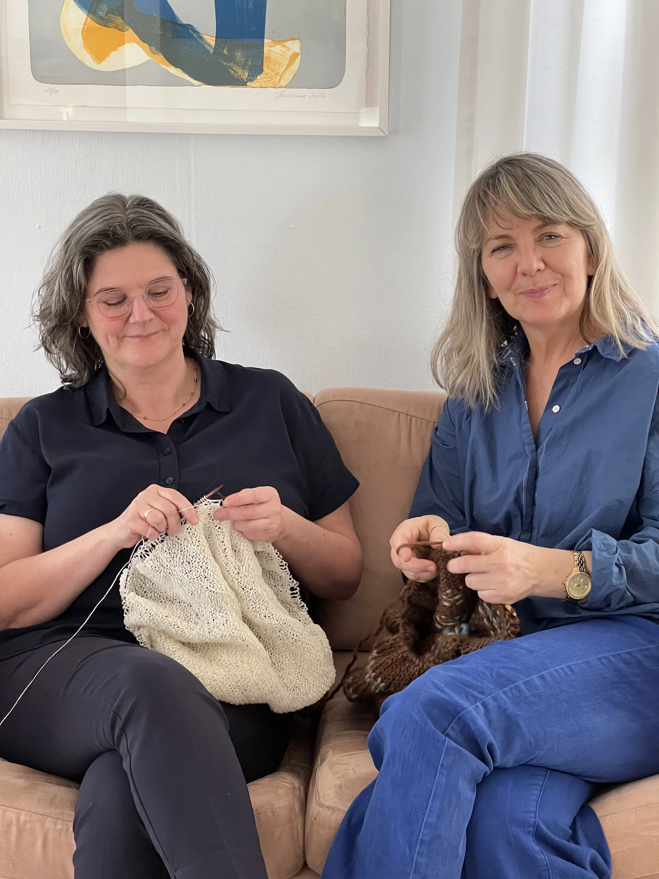 Two women sitting on a couch knitting, with one holding light yarn and the other holding dark yarn, in a cozy room with abstract art on the wall.