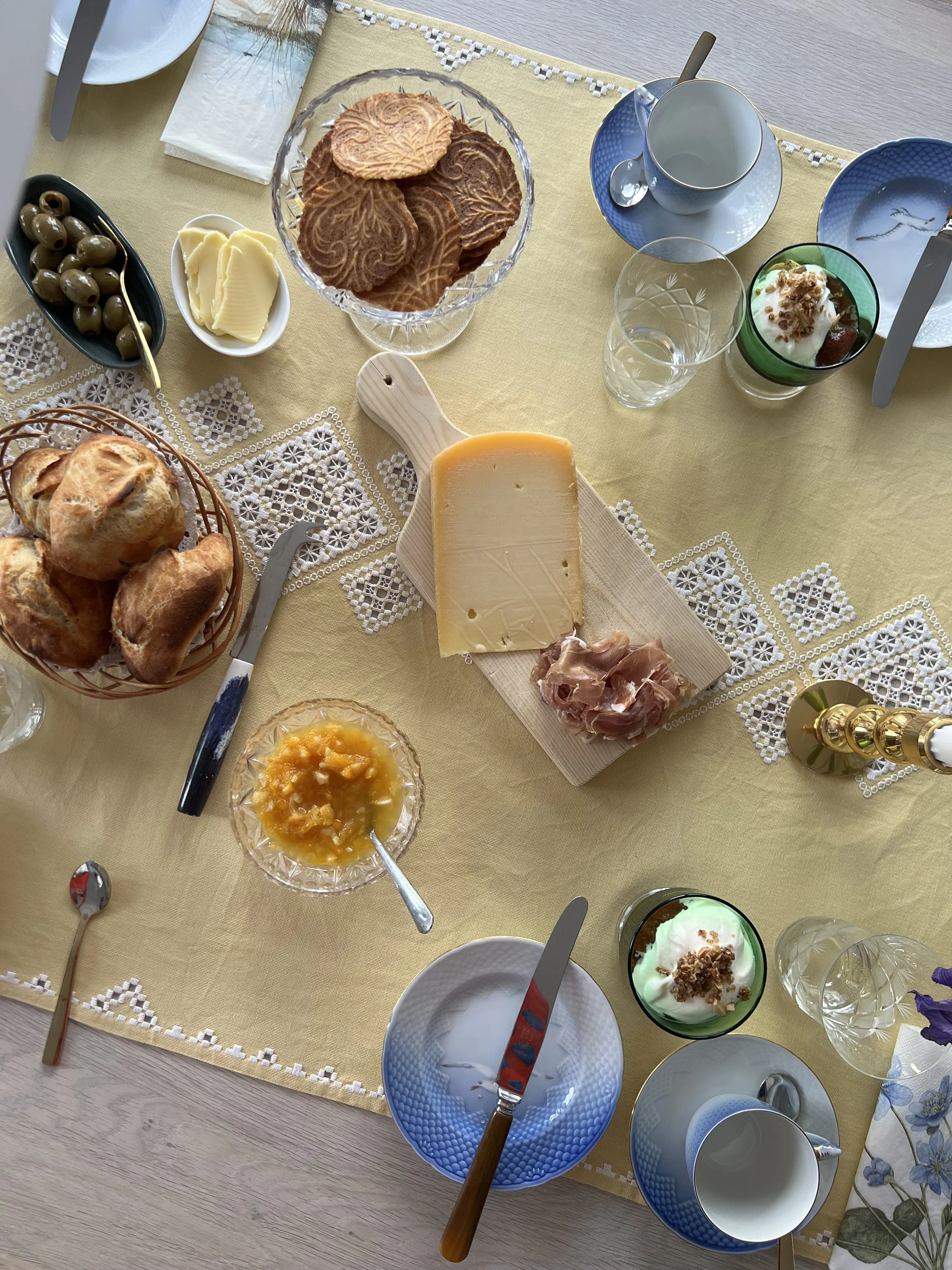 Breakfast table with rolls, cheese, butter, pastries, olives, jam, yogurt, and tableware on a yellow tablecloth. Tourists can get a taste of local delicacies when traveling in the Faroe Islands.