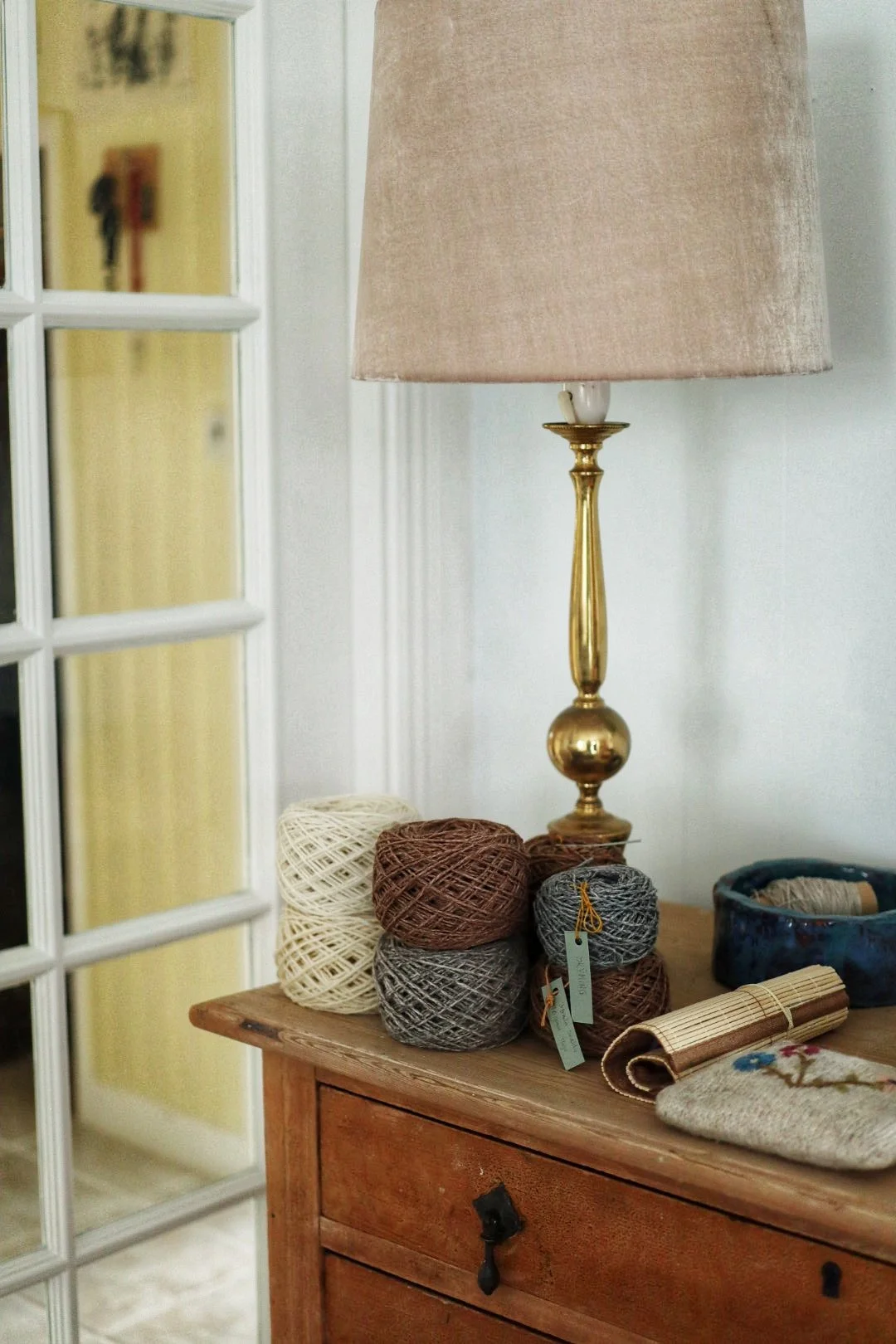 Wooden table with a brass lamp and stacked yarn balls in beige, brown, and gray colors, next to a blue bowl and a decorative pouch. Lamb wool.