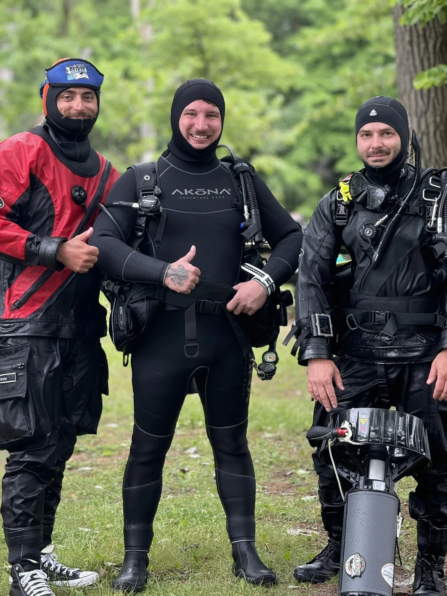 Three men in scuba diving gear, standing outdoors on grass with trees in the background. The man in the middle is giving a thumbs-up and smiling, while the other two are standing with relaxed expressions.