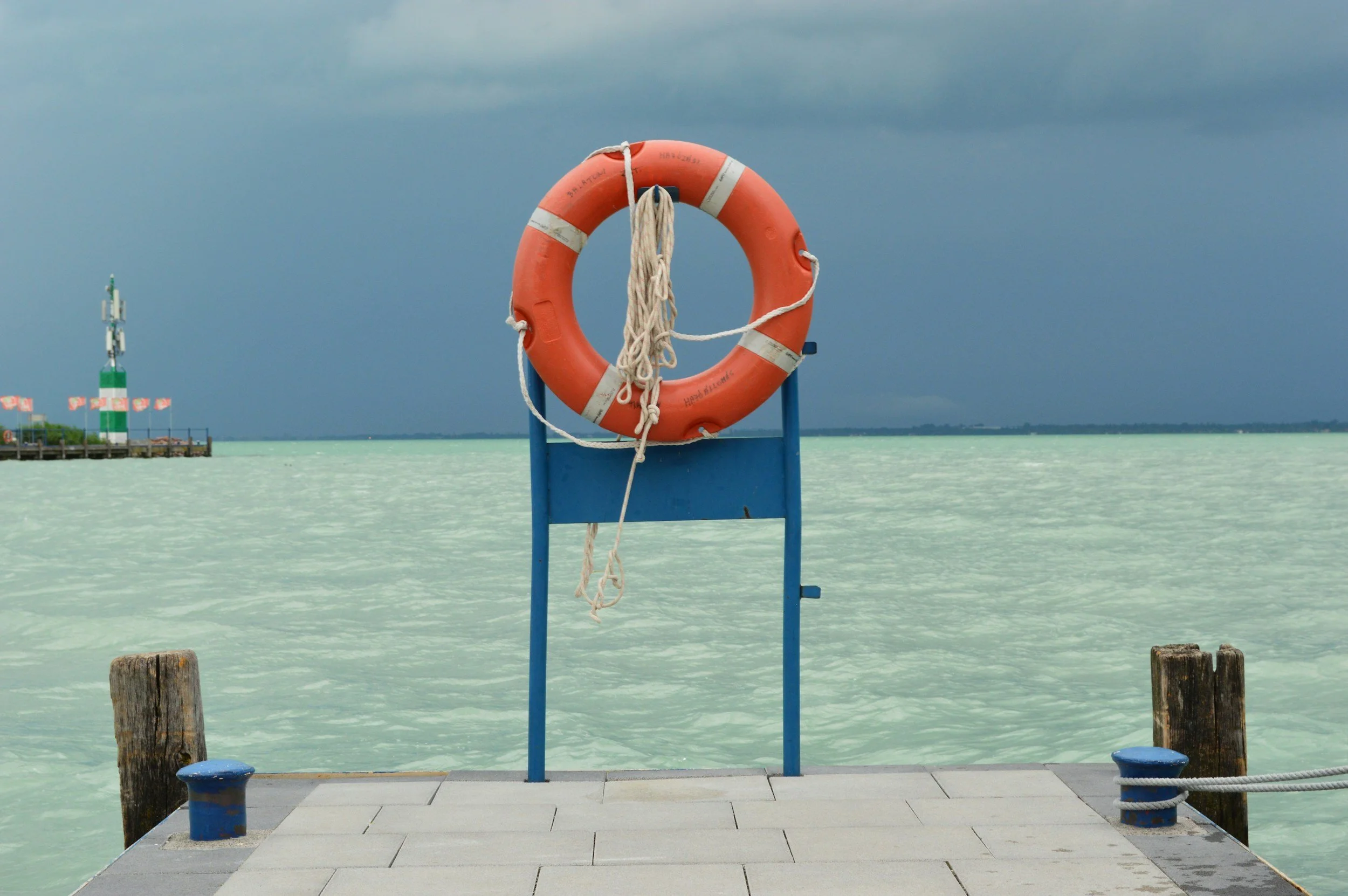 A lifebuoy on a blue metal stand on a dock by calm pale green water, near a weathered wooden post and a chain, with a distant lighthouse and cloudy sky.