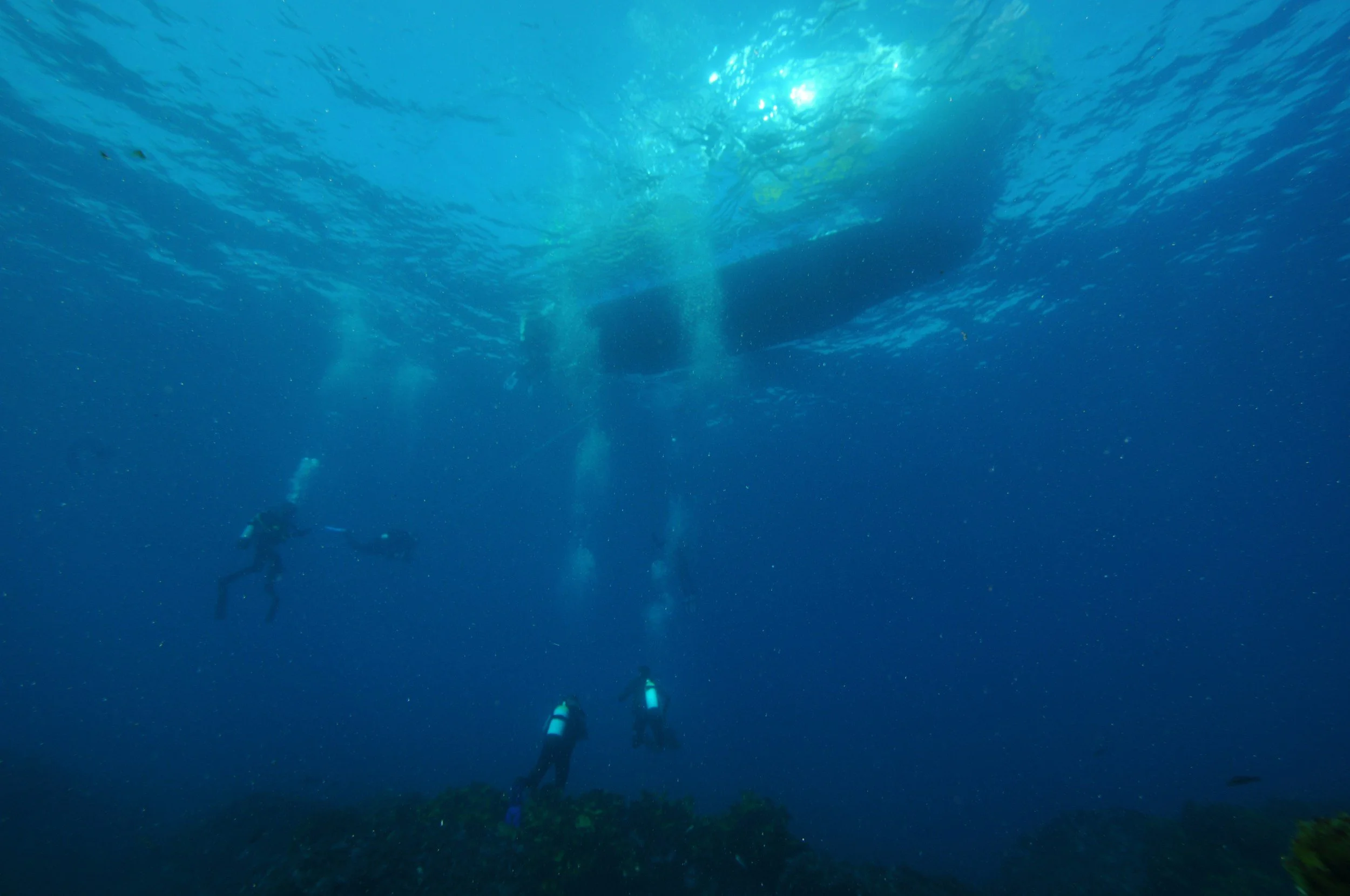 Three scuba divers exploring the ocean floor beneath a boat, with sunlight penetrating the water.