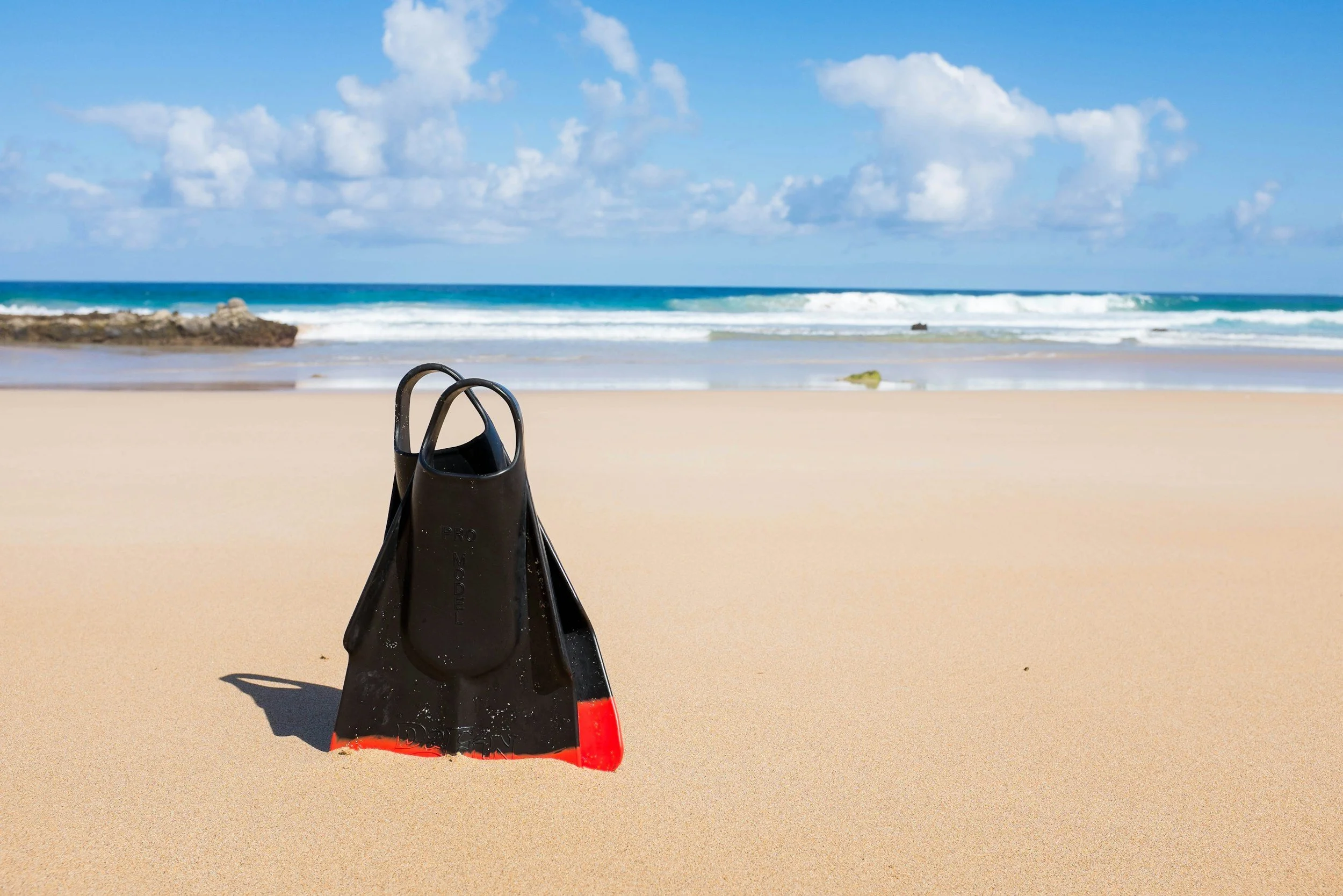 A pair of black snorkeling fins with a red tip stands upright in the sandy beach facing the ocean under cloudy blue sky.