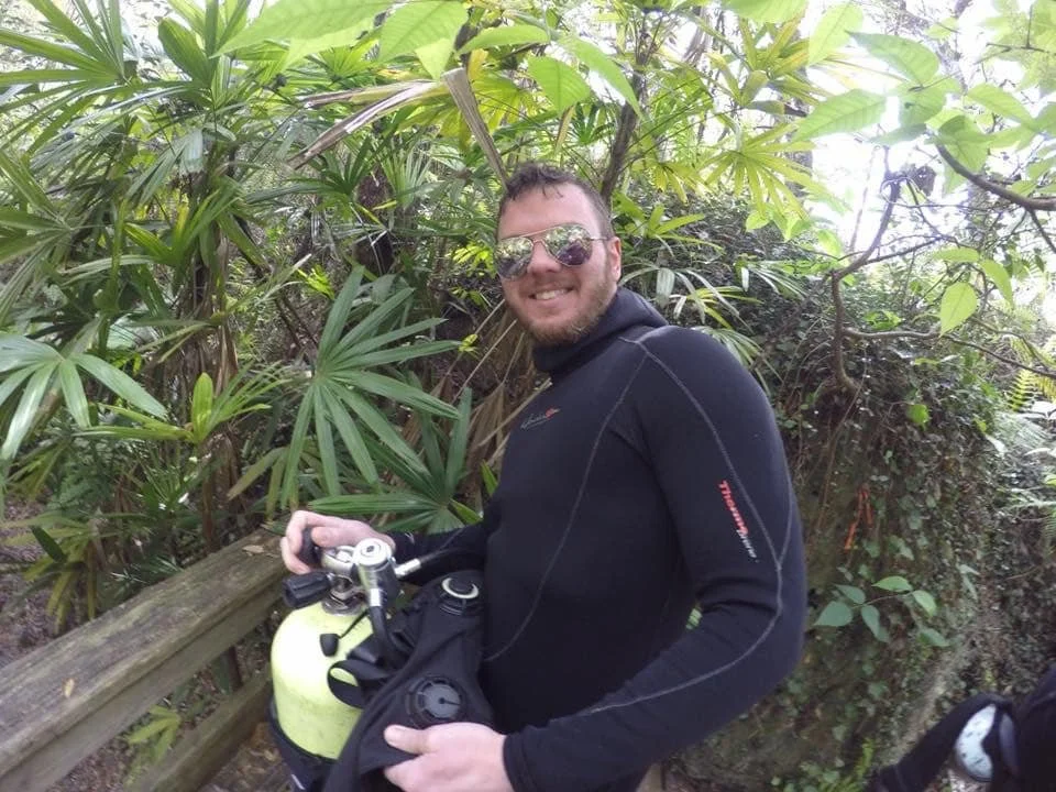 A man in black outdoor clothing and sunglasses standing on a wooden trail in a lush green forest, holding scuba equipment.