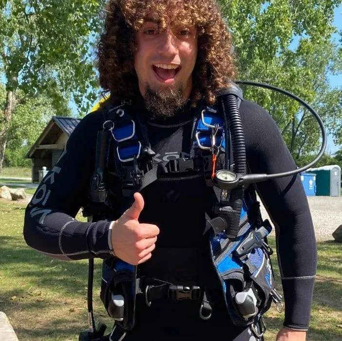 A man with curly hair and a beard outdoors, smiling and giving a thumbs-up while wearing scuba diving gear.