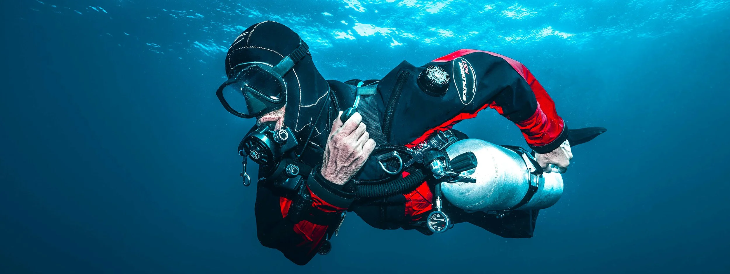 A scuba diver wearing a black mask and a black and red wetsuit, holding a diving tank, swimming underwater.