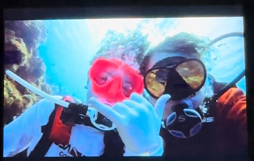 Two scuba divers underwater, one making an OK sign with their hand and the other wearing a red mask and blowing bubbles.