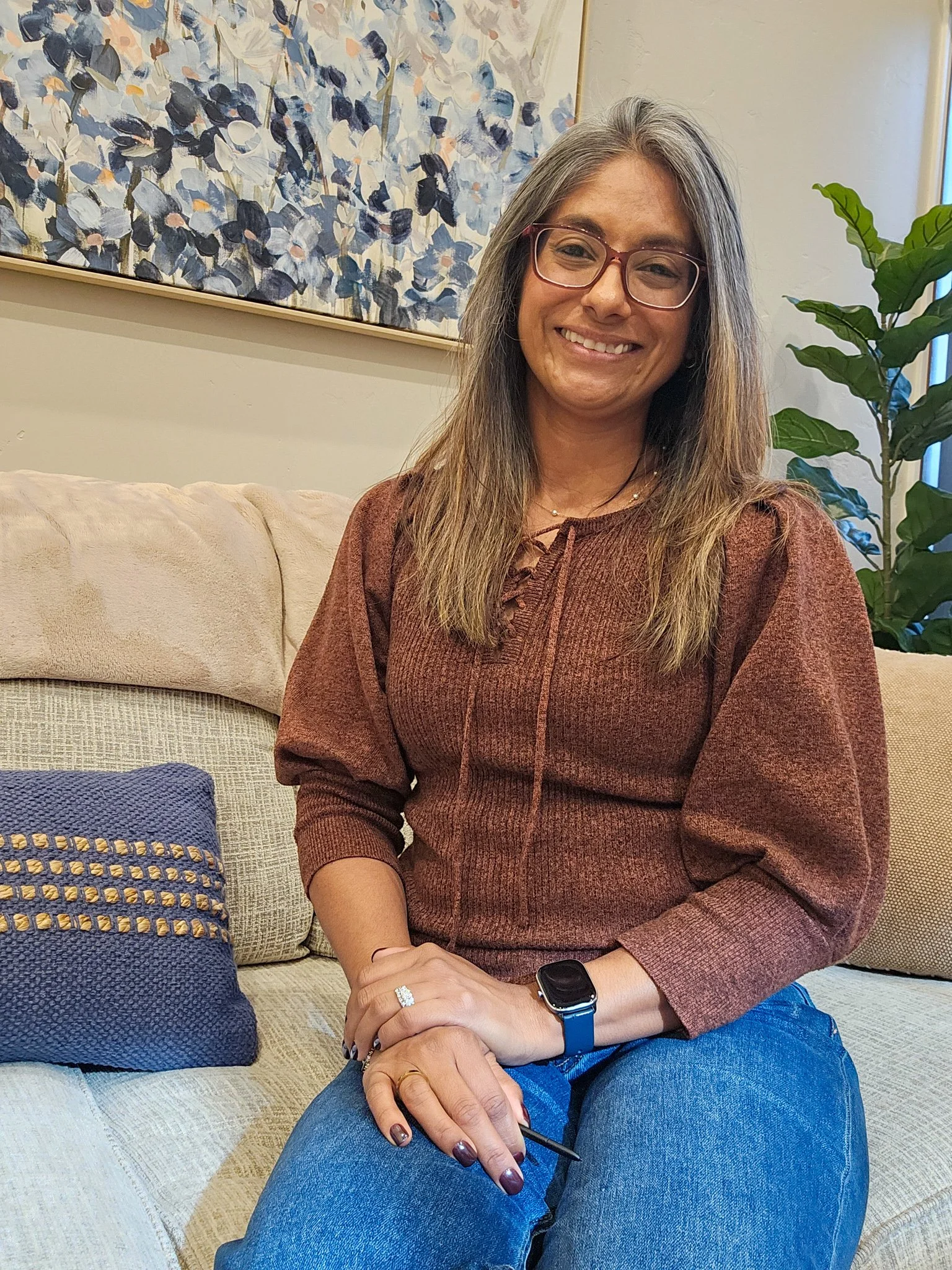 A woman with long gray hair wearing glasses, a brown sweater, and blue jeans, sitting on a beige couch with a patterned pillow, smiling at the camera, with a large abstract floral painting in shades of blue and white hanging on the wall behind her.