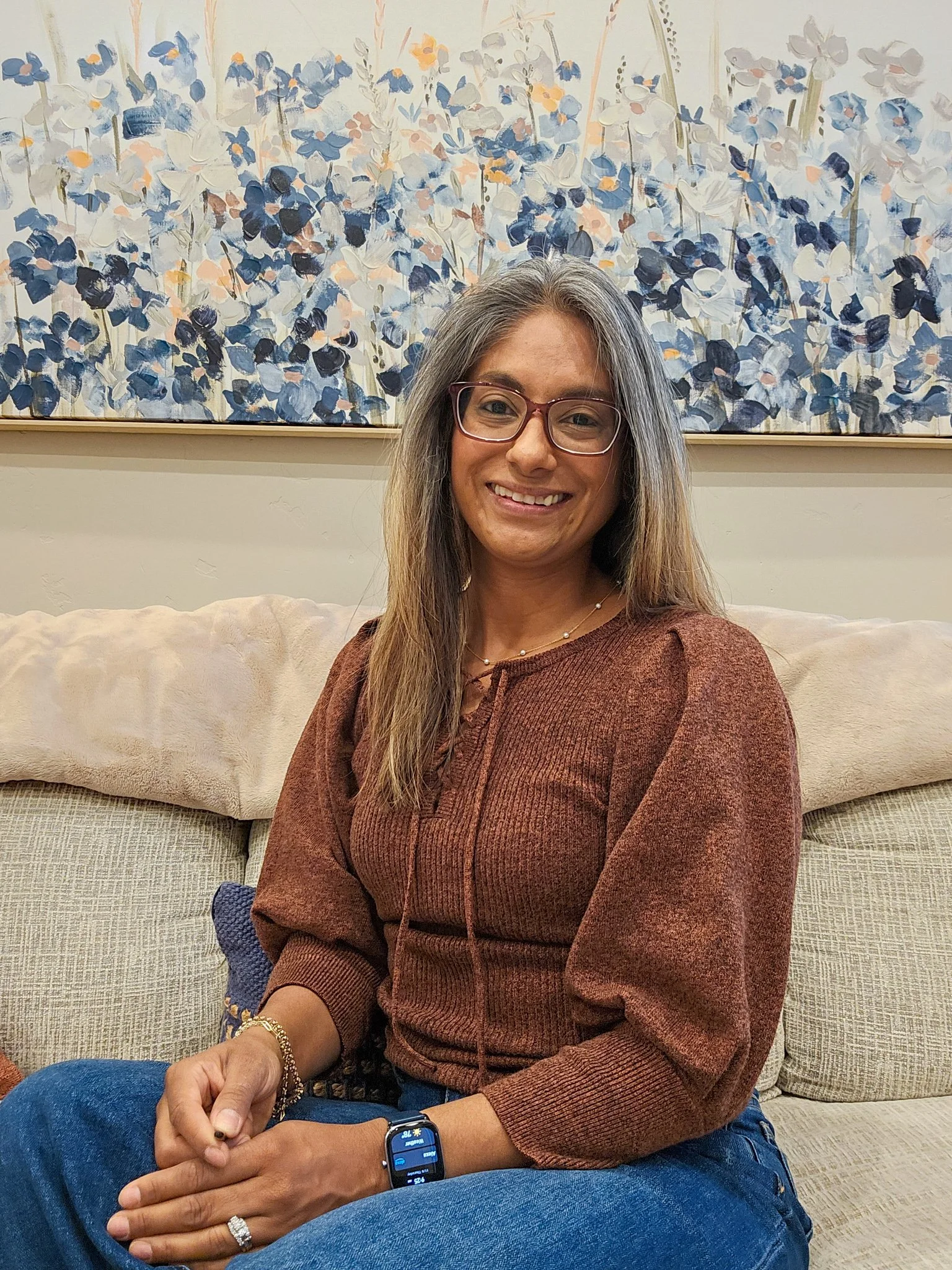 A woman with long gray hair, glasses, and a warm smile, seated on a beige couch in front of an abstract painting with blue, white, and yellow hues.