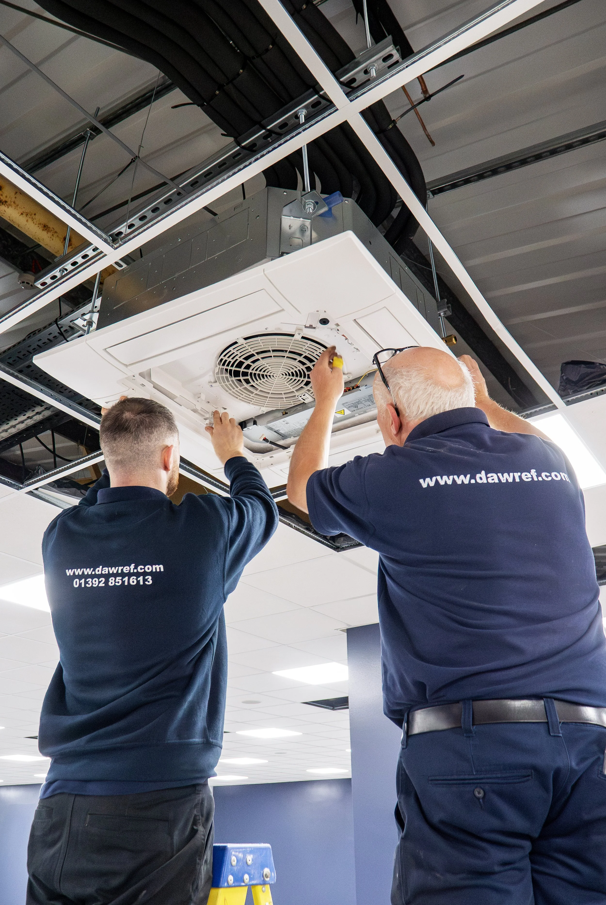 Two workers in navy blue uniforms installing or repairing an HVAC unit suspended from a ceiling.