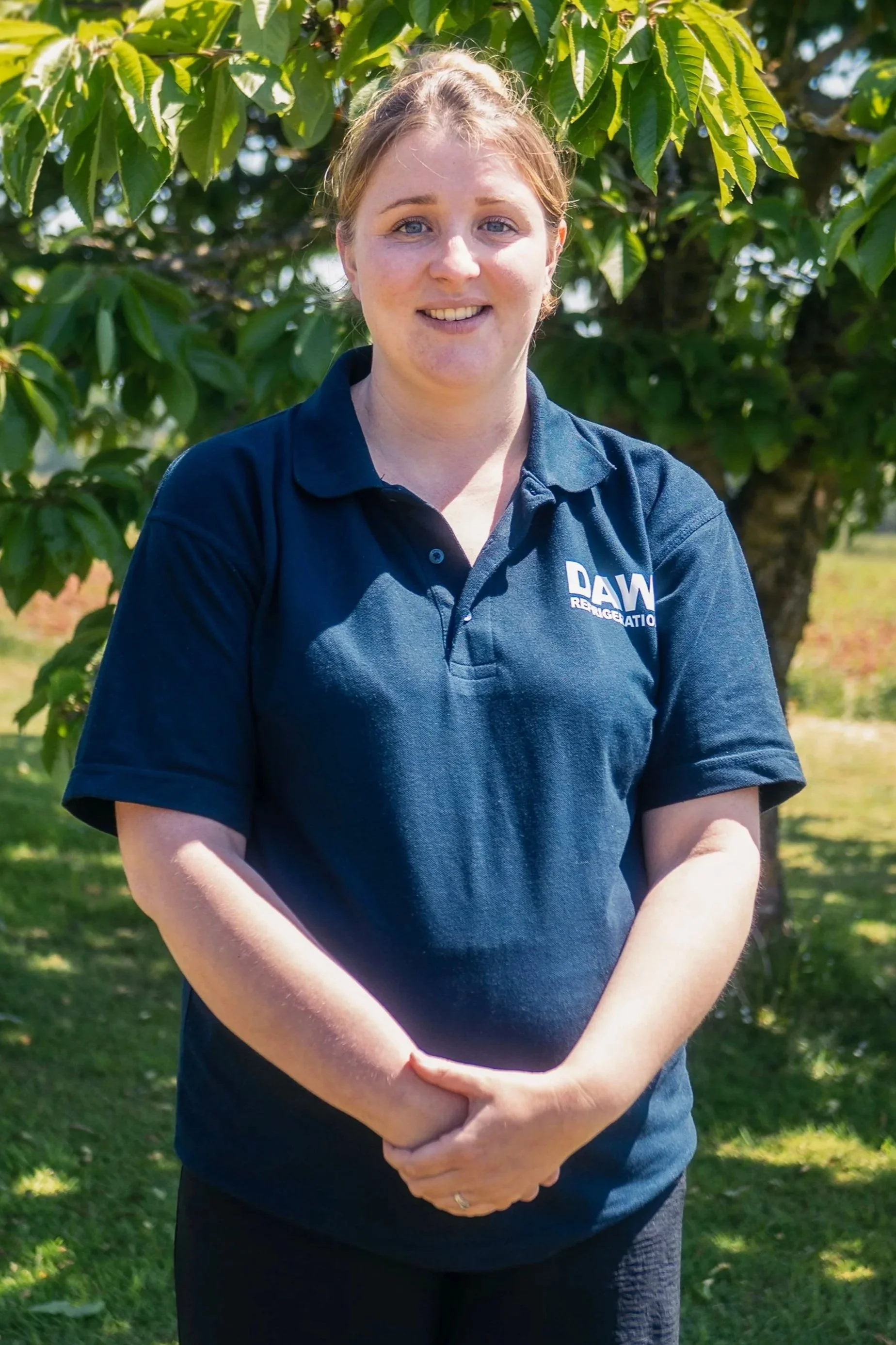 A woman with blonde hair and blue eyes is standing outdoors in front of a leafy tree, smiling. She is wearing a navy blue polo shirt with a logo that says "DAW Refrigeration" on the chest, and has her hands clasped in front of her.