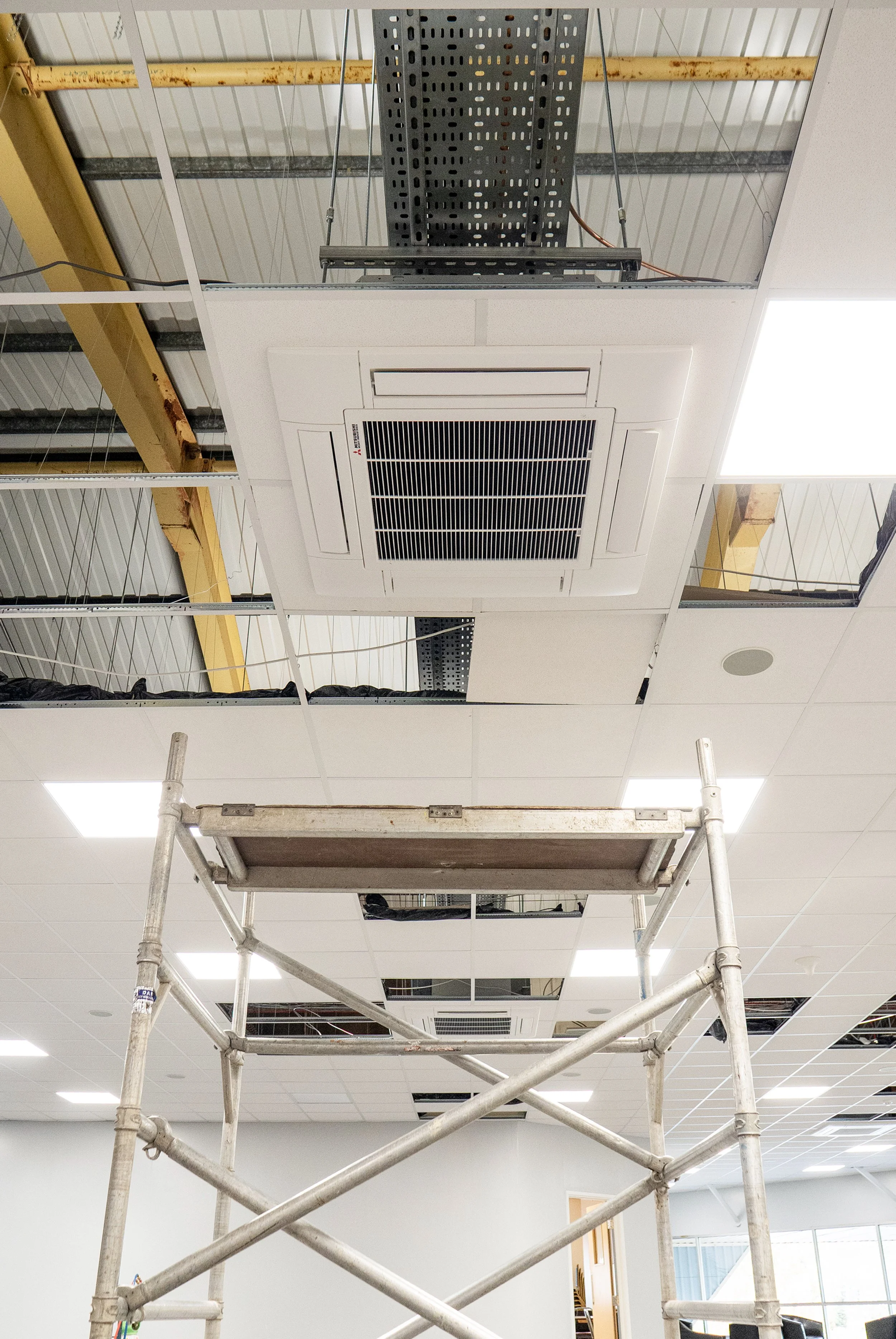 The ceiling of an indoor space under construction showing an installed white ceiling air conditioning vent. There are yellow and black metal beams, scaffolding, and partially removed ceiling tiles. Some wiring and metal framework are visible.