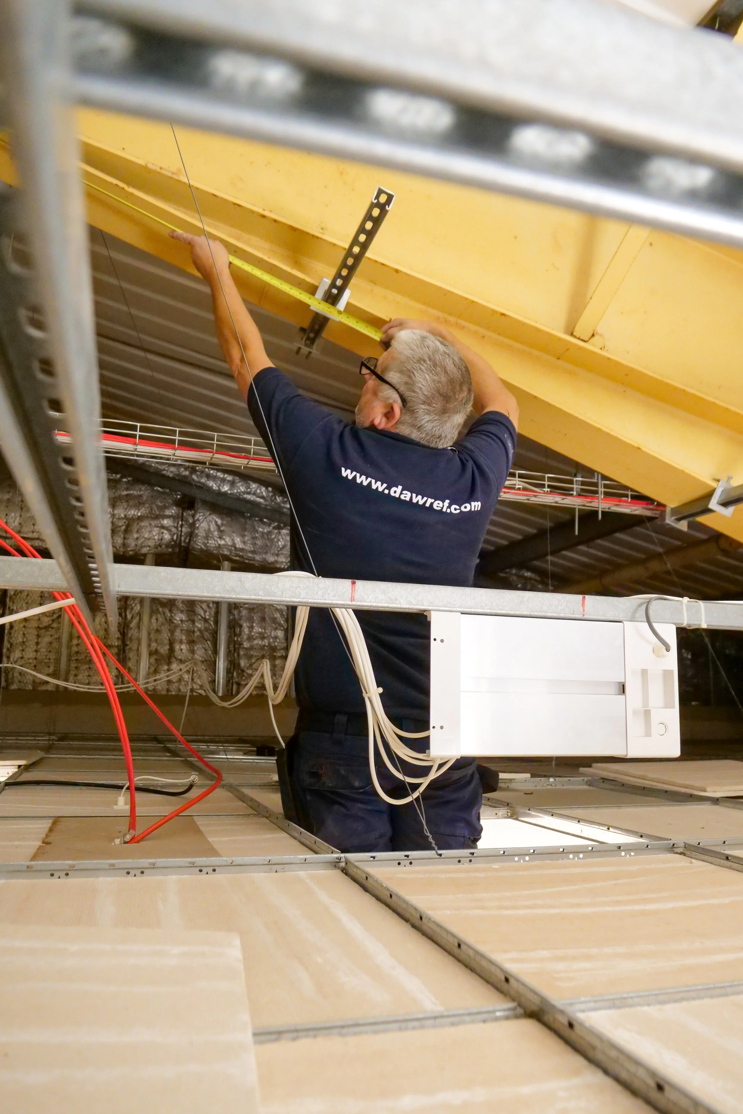 A man wearing a navy blue shirt with a website URL on the back is working in an industrial setting. He appears to be taking measurements with a yellow tape measure for installing or repairing equipment. The environment includes metal scaffolding, wir