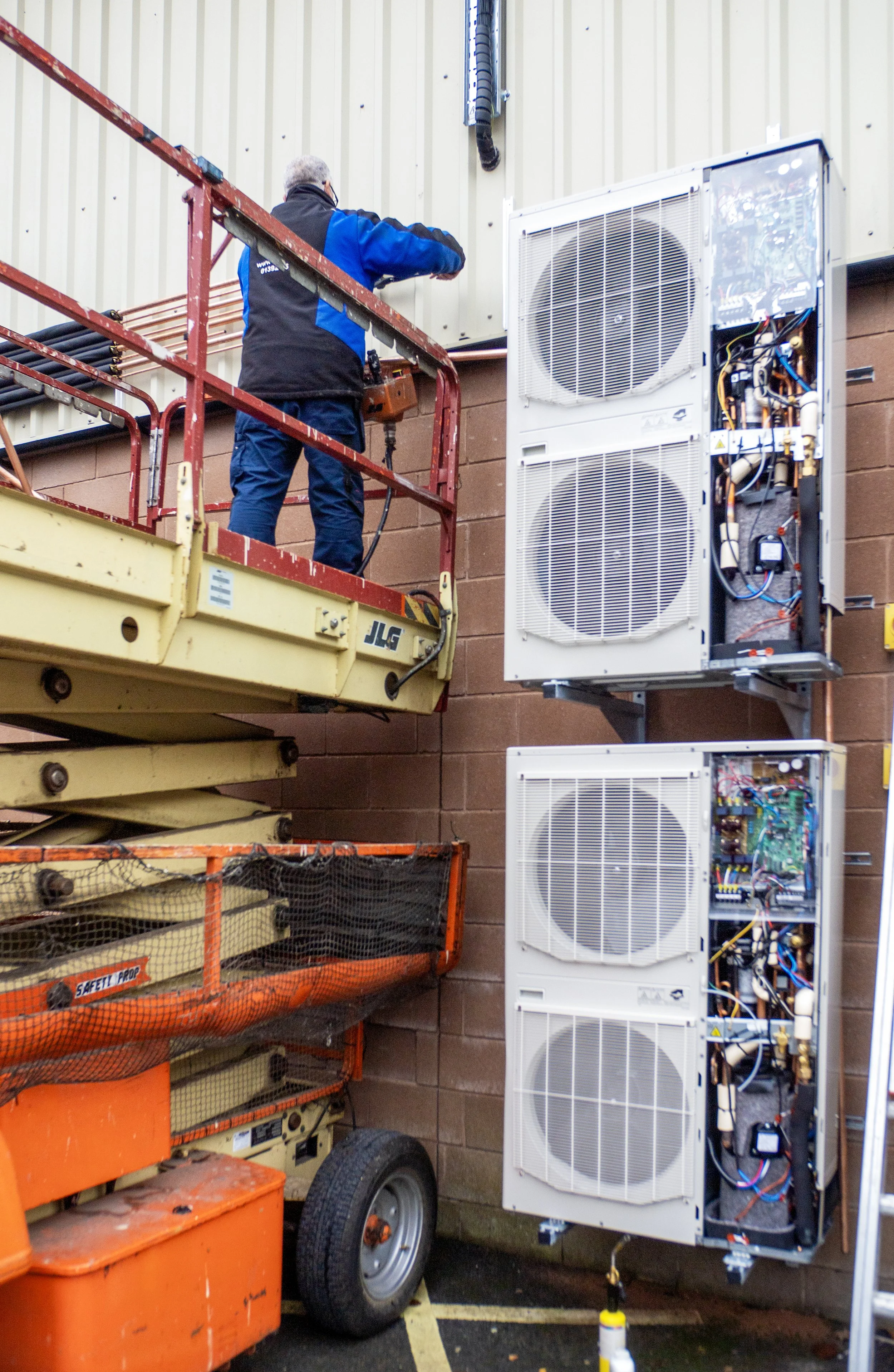 A man working on HVAC equipment, standing on a yellow scissor lift, with HVAC units and tools around in a parking lot.