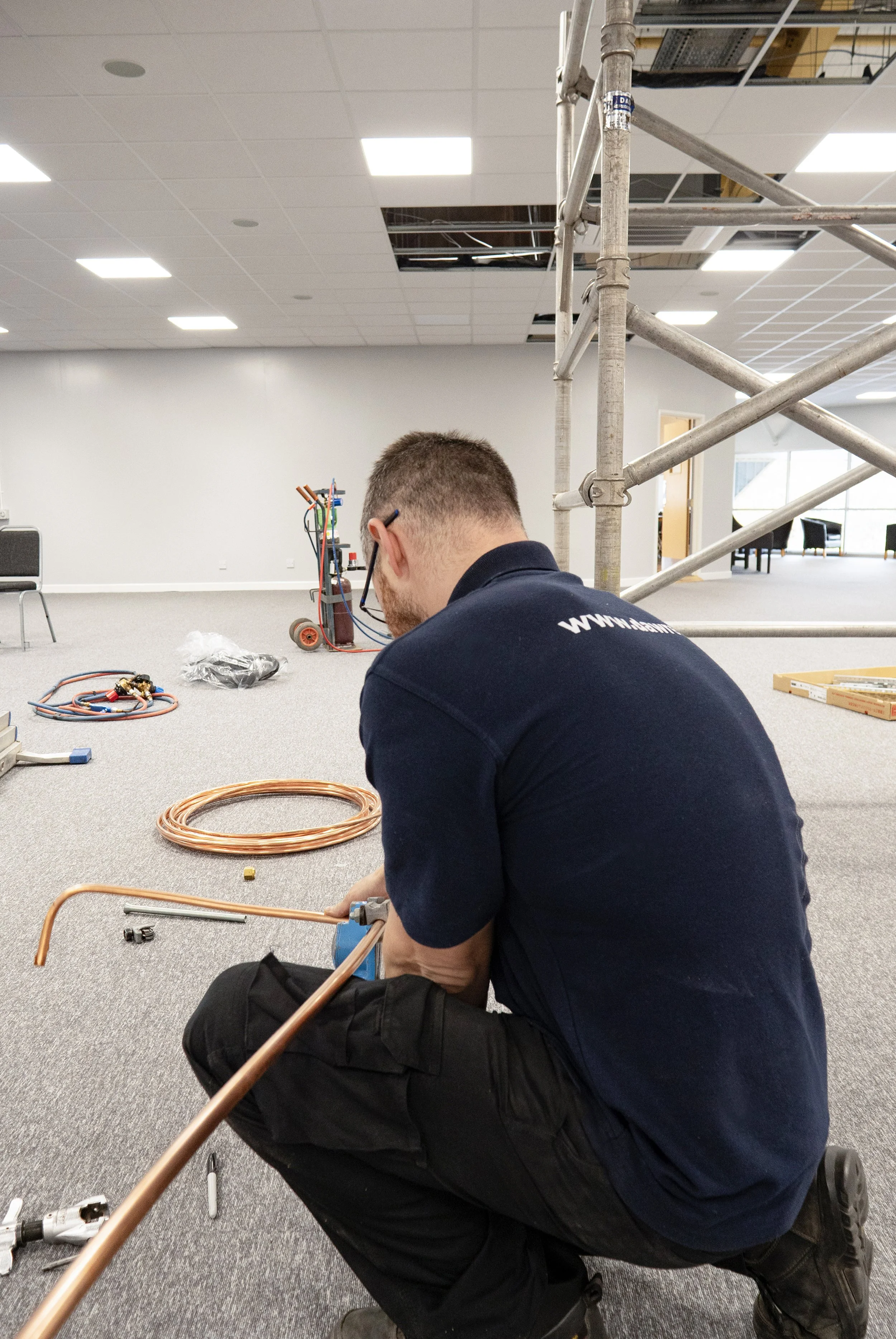 A technician kneels on the floor working with electrical wires and tools, with a large metal scaffold and open ceiling panels in the background.