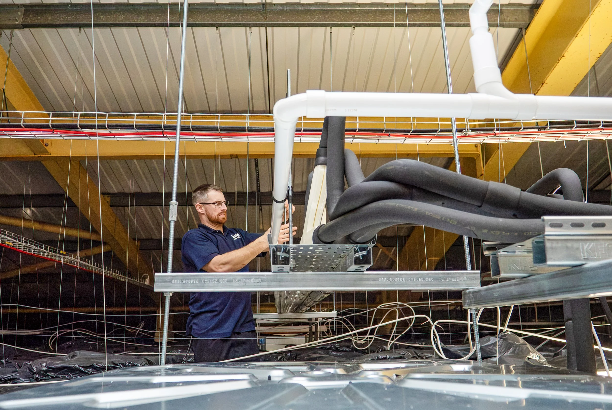 A man wearing glasses and a navy blue polo shirt is working with electronic equipment and cables in an industrial setting with yellow beams and a metal roof.