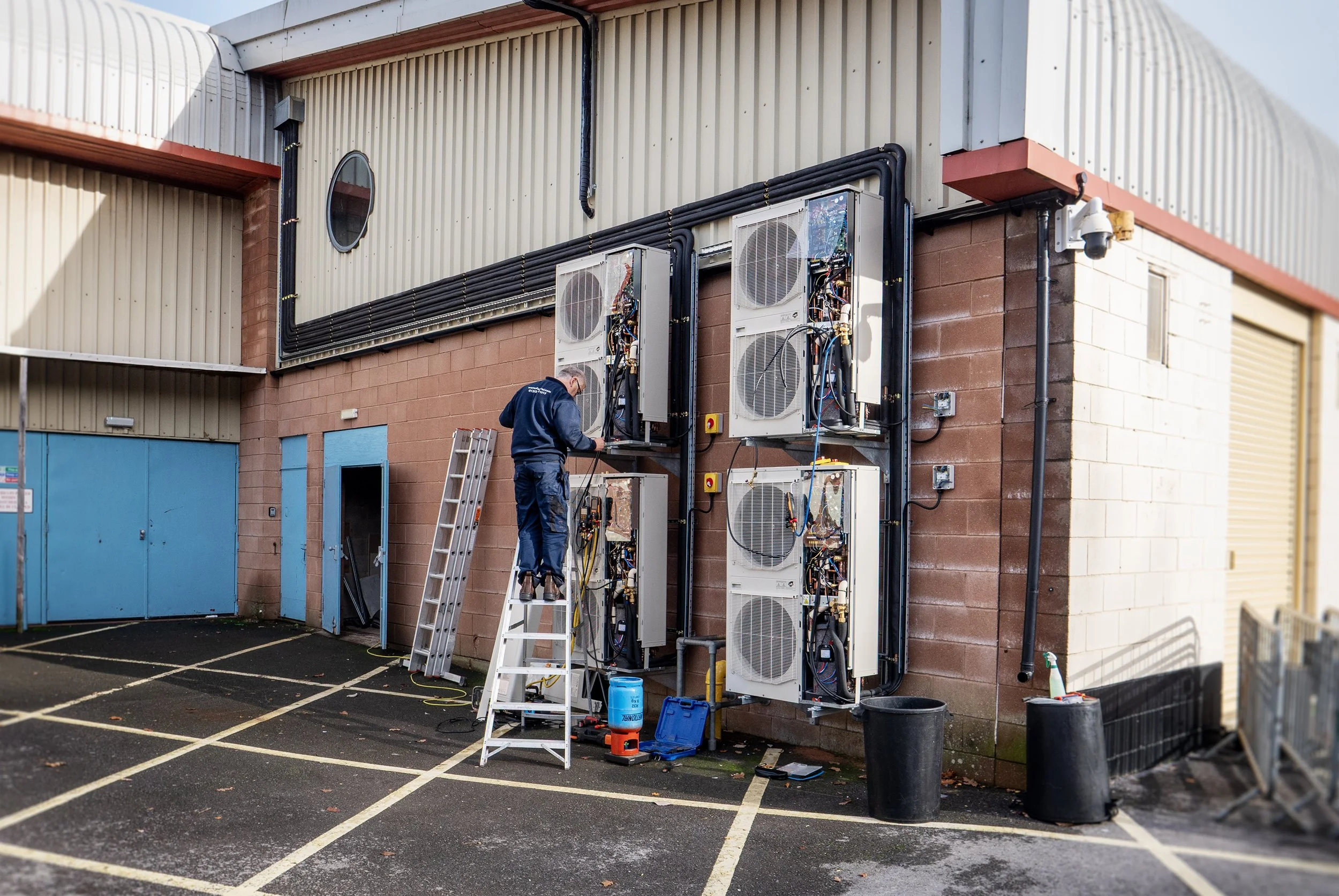 A technician working on an outdoor HVAC or refrigeration unit, with tools and equipment around him, outside a commercial building.
