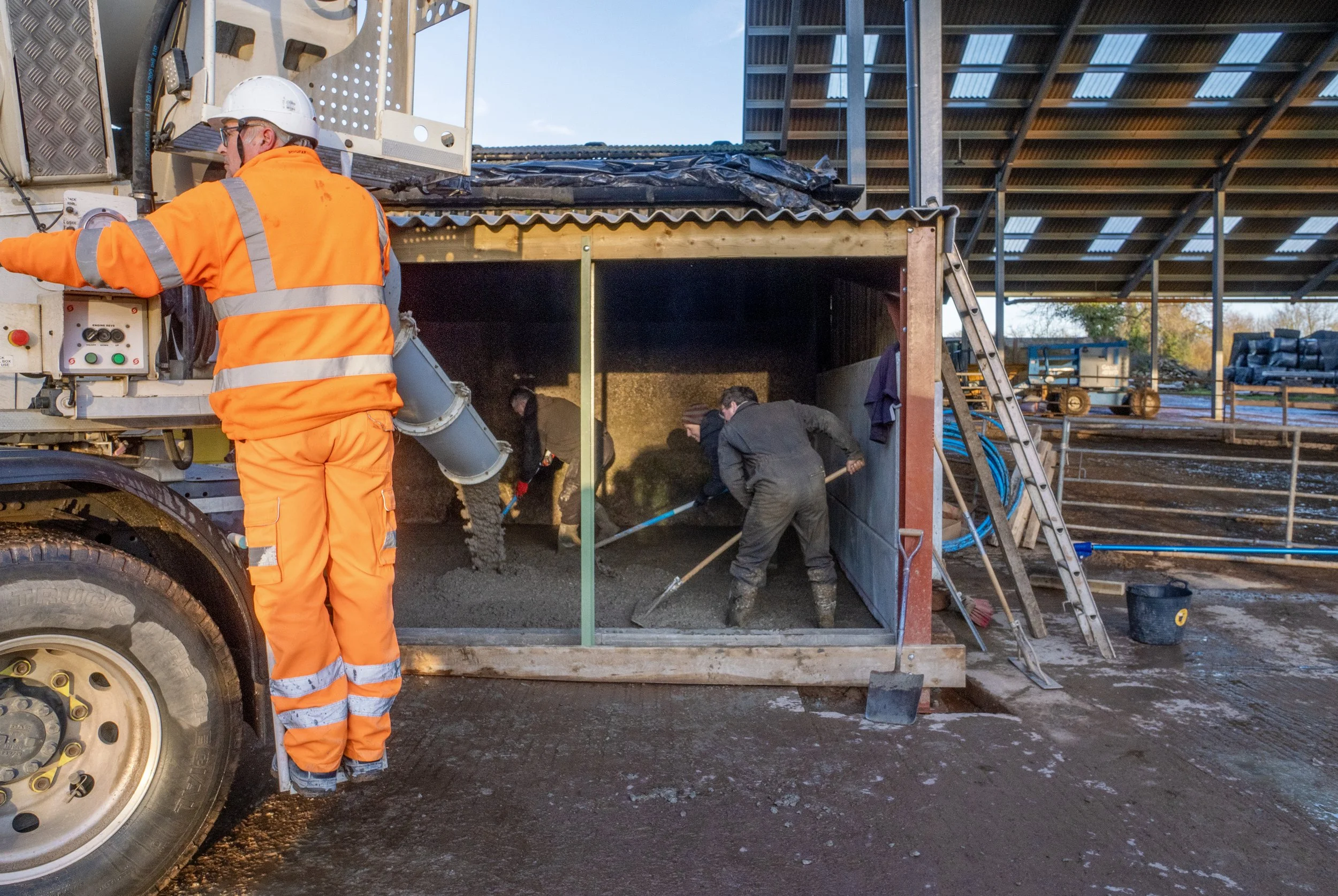 Workers in safety gear spreading concrete at a construction site under a metal roof.
