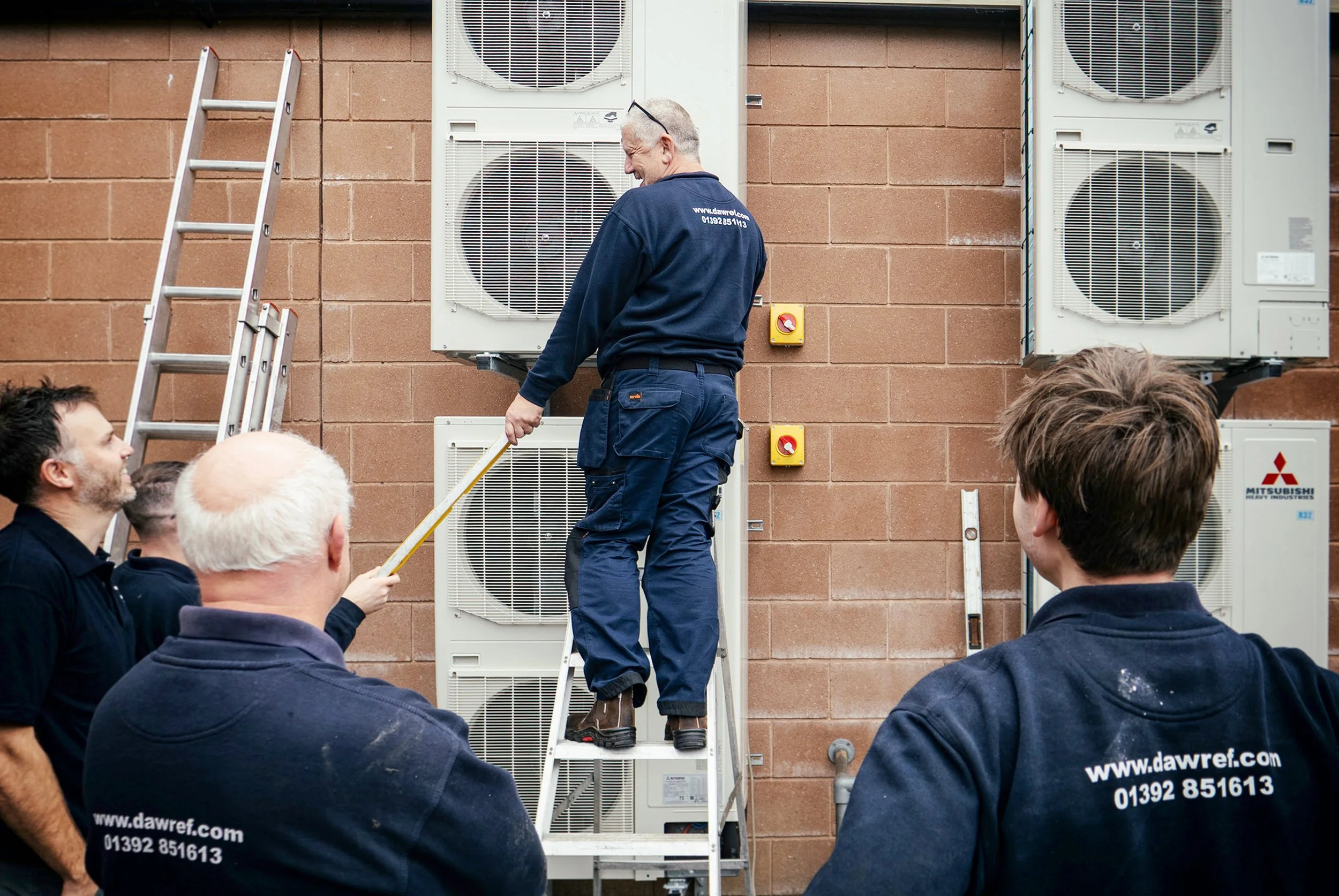 A group of men gathered outdoors watching a technician demonstrating or repairing air conditioning units mounted on a brick wall. The technician is standing on a ladder, wearing a navy uniform with the website 'www.dawref.com' and phone number visibl