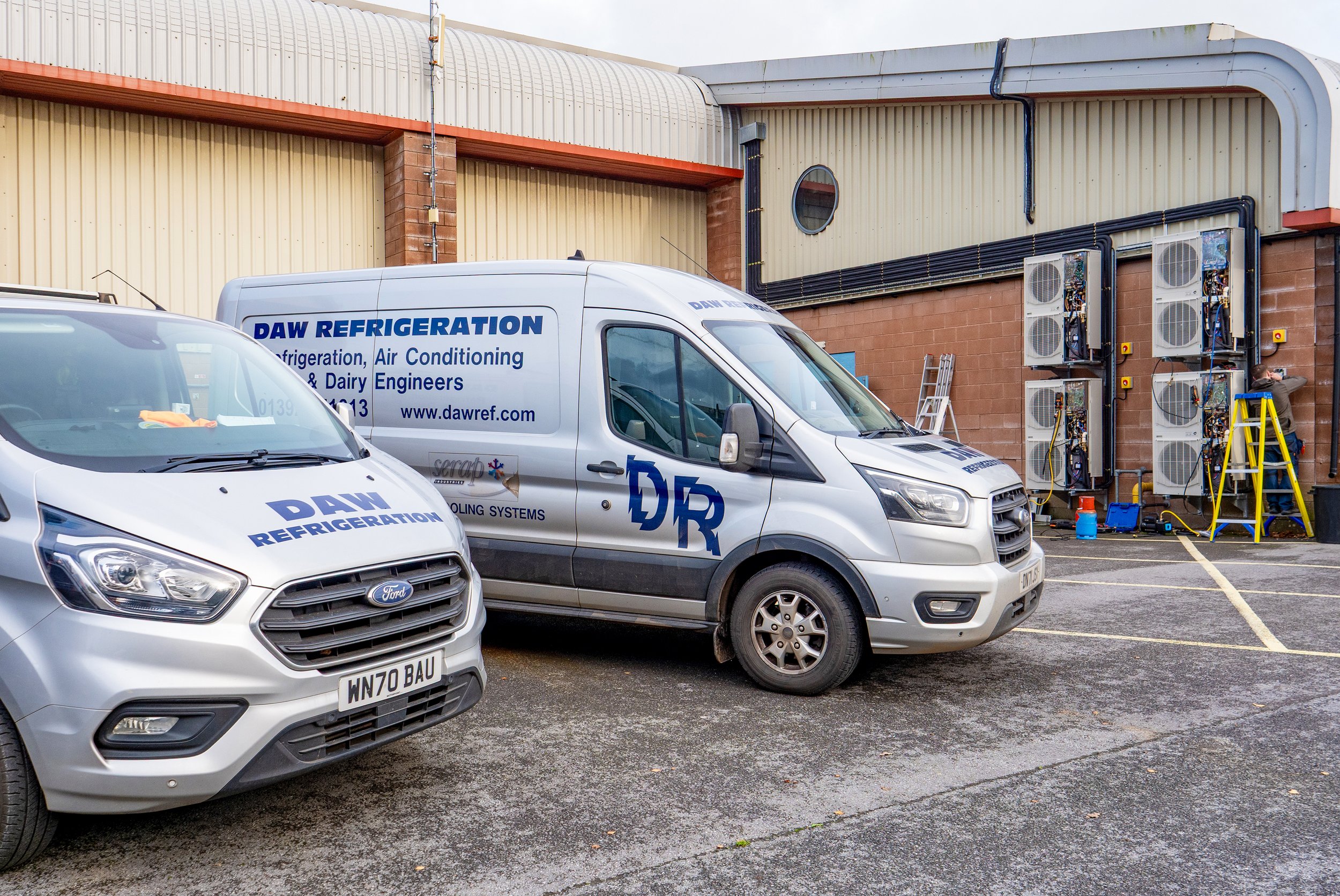 Two white service vans with blue text and logos parked outside a building with air conditioning units and electrical panels, with a worker installing or repairing equipment on the wall.