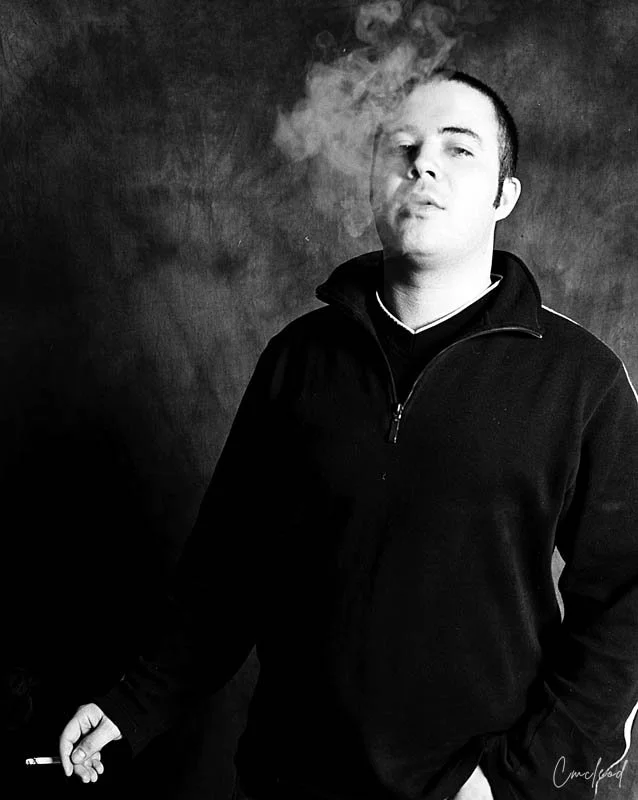 Black and white photo of a man in a dark zip-up jacket with short hair, holding a cigarette in his left hand, with smoke rising near his face, against a plain dark background.