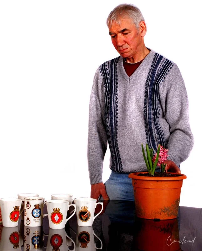 An elderly man with gray hair, wearing a patterned sweater, looking down at a potted plant with pink flowers on a table. Several white coffee mugs with colorful symbols are arranged on the table.