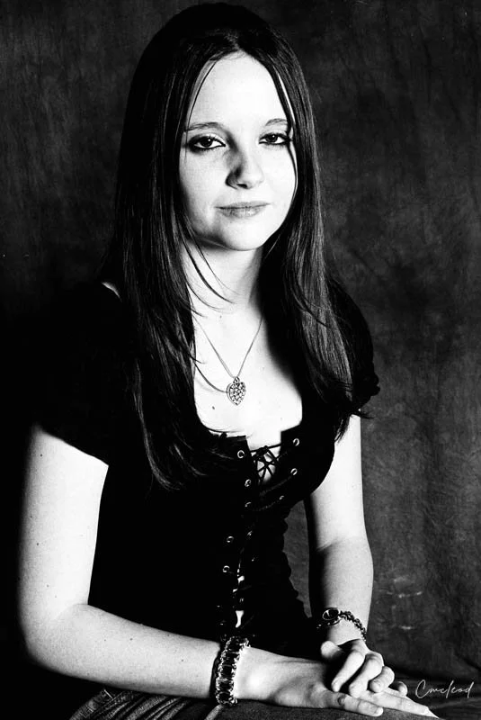 A young woman with long dark hair, wearing a black lace-up top, a pendant necklace, and bracelets on her left wrist, sitting against a dark background. The image is in black and white.