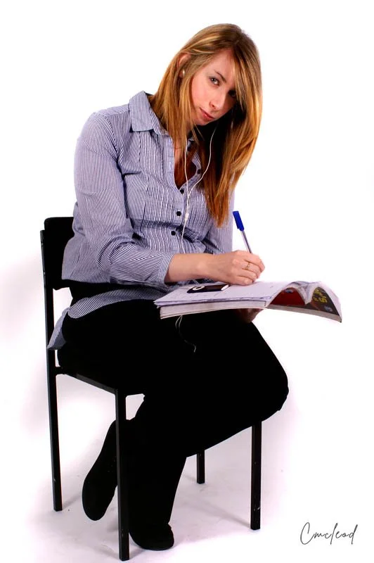 A woman sitting on a black chair, looking at a magazine, writing with a pen, listening to earphones, wearing a striped shirt and black pants, against a white background.