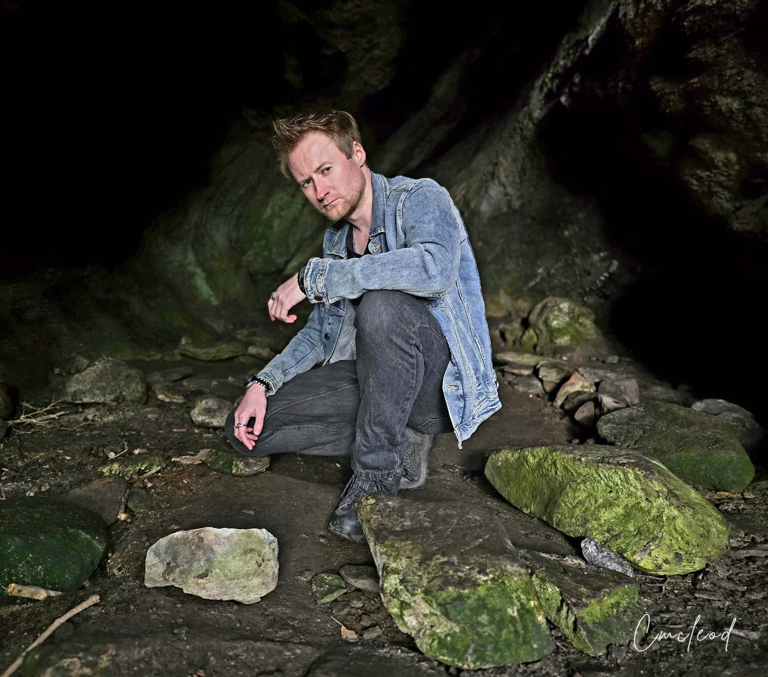 A man with light brown hair and a beard, wearing a denim jacket, black jeans, and black shoes, is kneeling on rocks with moss in a dark cave or tunnel setting.