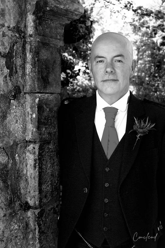 Black and white portrait of a bald man in a suit, standing next to a stone wall outdoors.