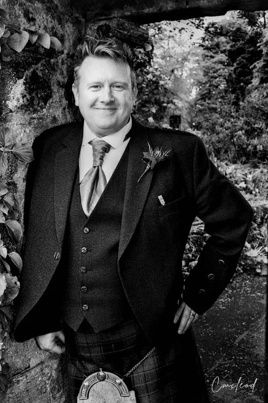 Black and white photo of a man in traditional Scottish attire, including a kilt and sporran, standing outdoors near a stone wall with trees in the background.