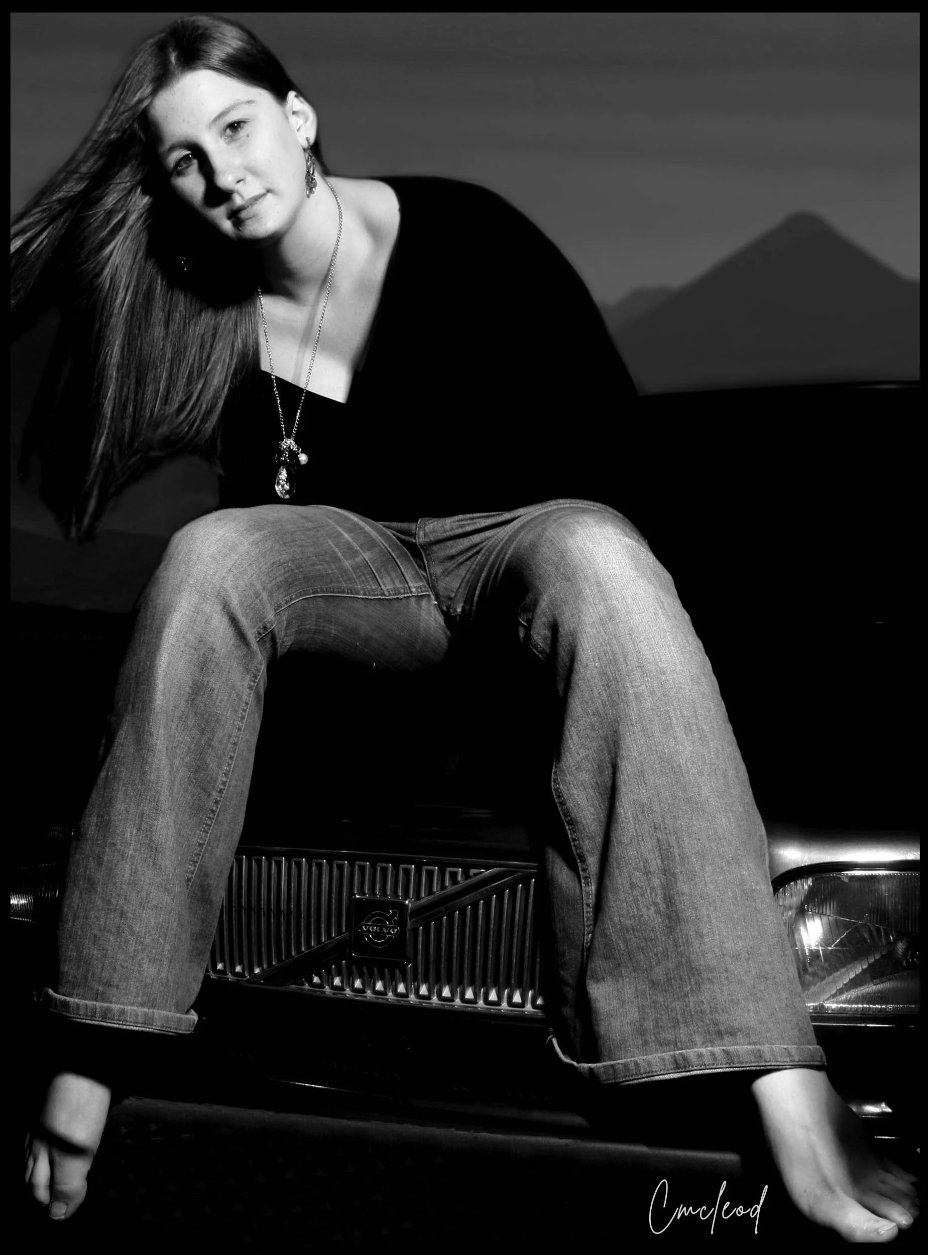 Black and white portrait of a young woman with long hair, sitting on the hood of a vintage Volvo car, wearing jeans and a black top, with a necklace and earrings, against a background of mountains.