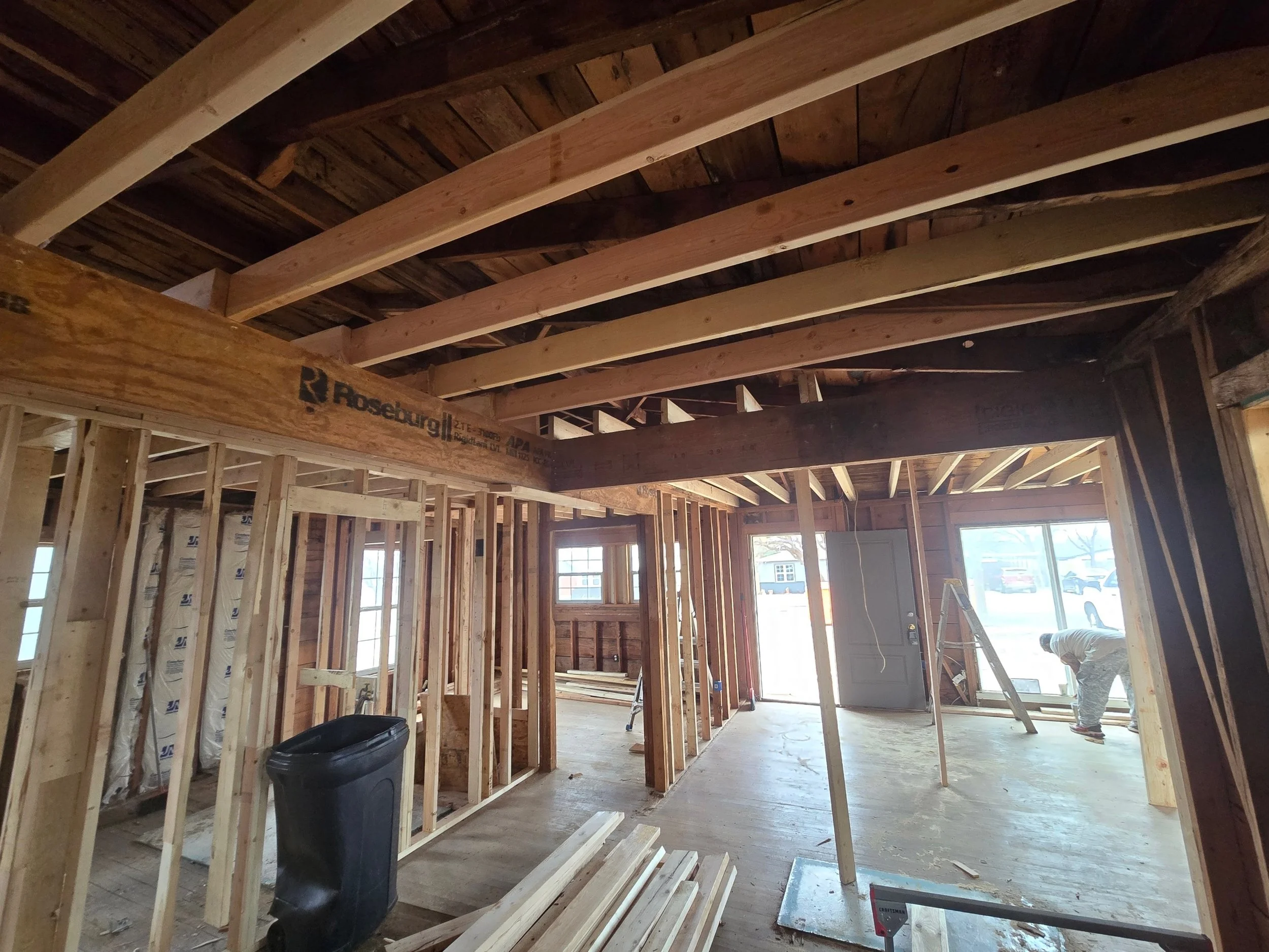 Interior view of a house under construction with exposed wooden framing, an open doorway, large windows, and a construction worker working near the front door.