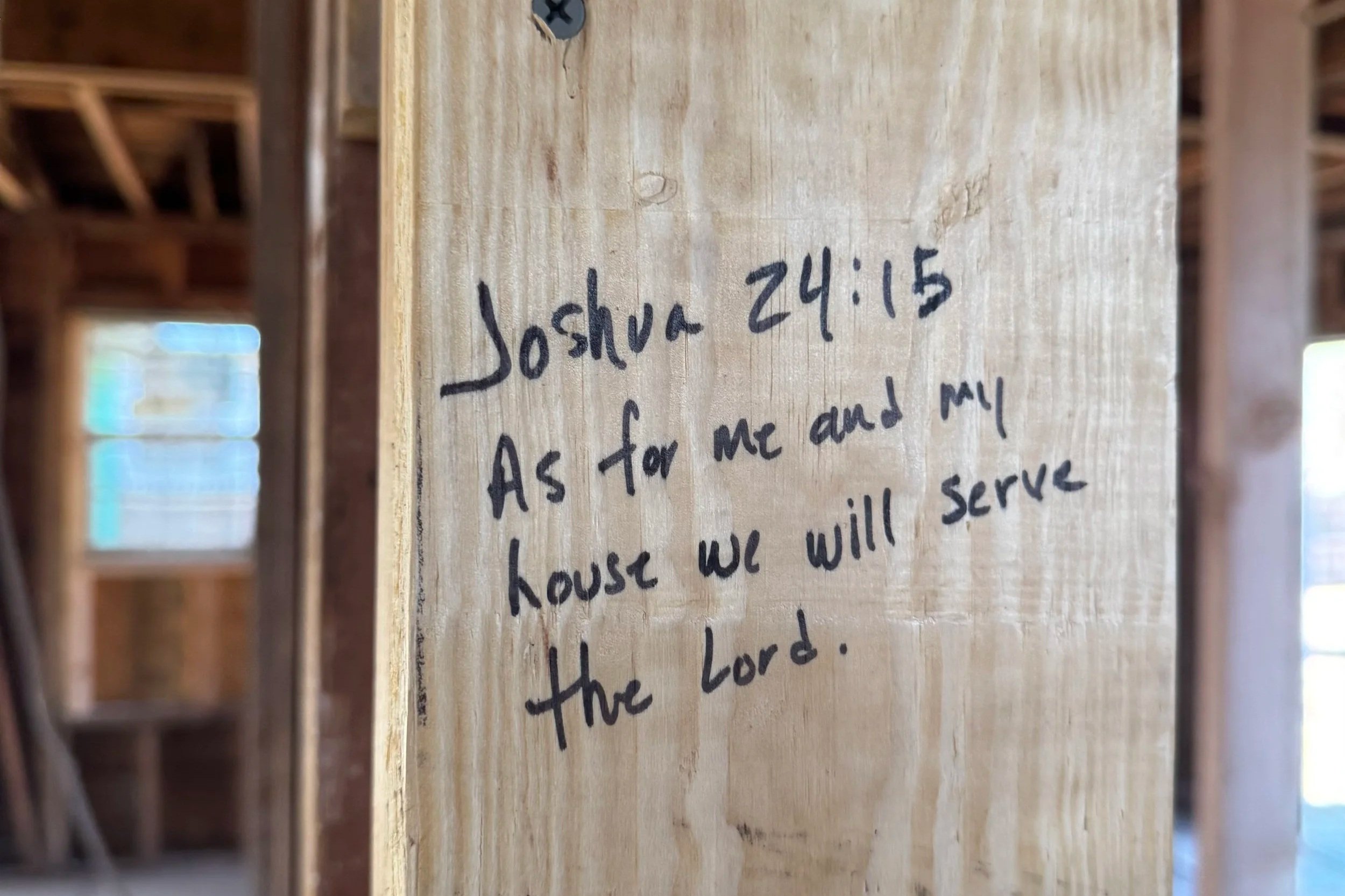A wooden beam inside a house under construction with a handwritten Bible verse, Joshua 24:15, about serving the Lord.