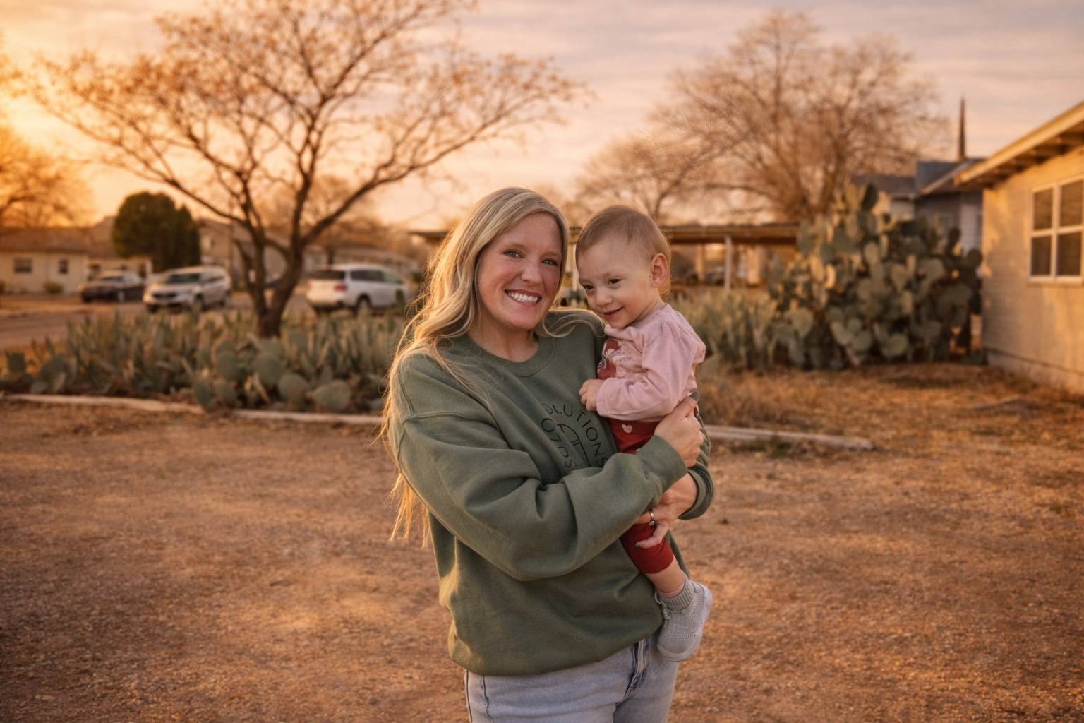 A woman holding a smiling baby outdoors during sunset, with trees, a house, cacti, and parked cars in the background.